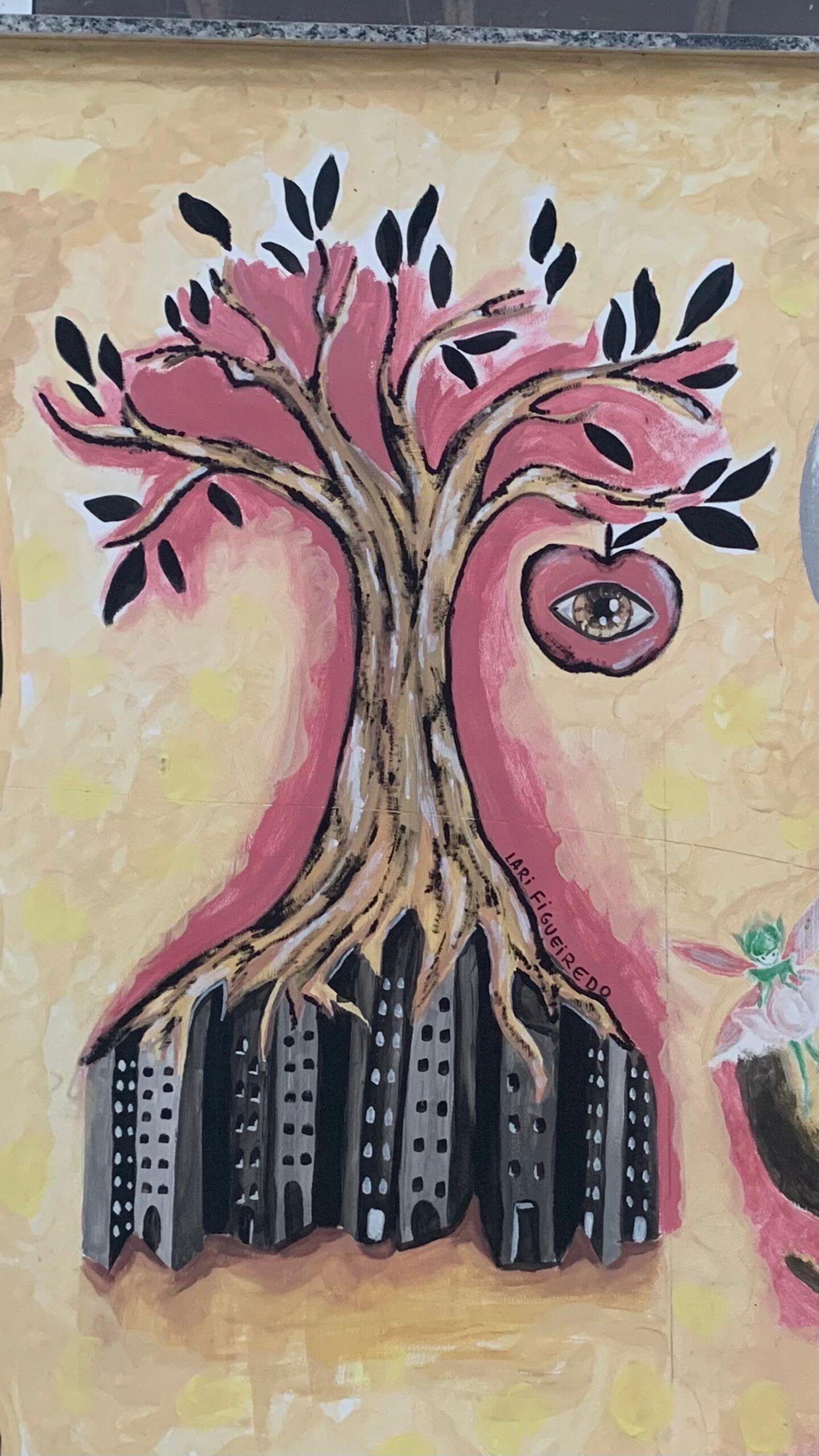 Pintura em uma parede com fundo em tons de amarelo e bege. A imagem central é uma árvore estilizada com raízes profundas, mas incomuns: o tronco marrom e os galhos se abrem em um halo rosa-avermelhado, e as raízes se ramificam sobre e dentro da silhueta escura de uma paisagem urbana, com prédios cinza-escuros no lugar do solo. As folhas da árvore são pretas. Pendurada em um dos galhos, há uma maçã vermelha com um olho humano pintado em seu centro.