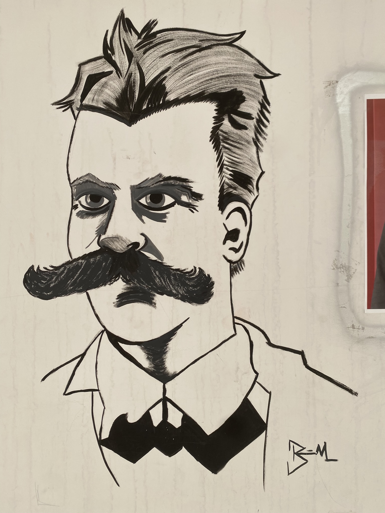 Desenho em preto e branco em uma parede clara, de um busto masculino em estilo de caricatura. O homem tem um cabelo espetado e escuro, sobrancelhas grossas e um bigode longo e escuro, com expressão séria ou intensa. Ele está vestido com uma camisa de colarinho e uma gravata borboleta escura.