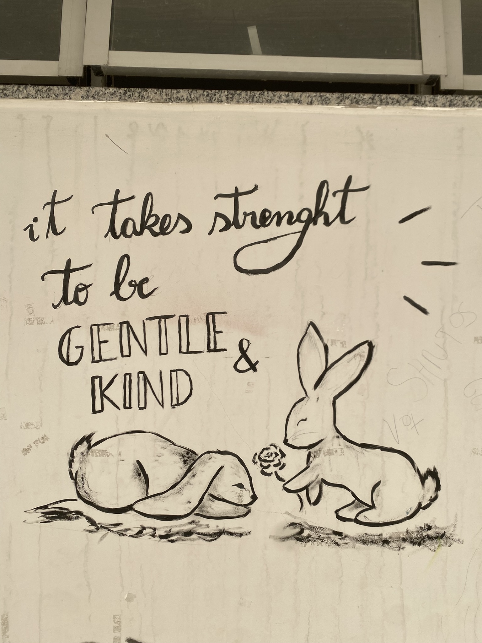 Desenho a caneta preta em uma parede clara, que ilustra a frase "it takes strenght to be GENTLE & KIND" em caligrafia cursiva e em caixa alta. Abaixo da frase, dois coelhos estão desenhados no chão: um está deitado com os olhos fechados, e o outro está sentado de frente para ele, segurando uma pequena rosa na boca. O desenho dos coelhos e o texto são em preto, com traços de realce ao redor da frase e da cabeça do coelho.
