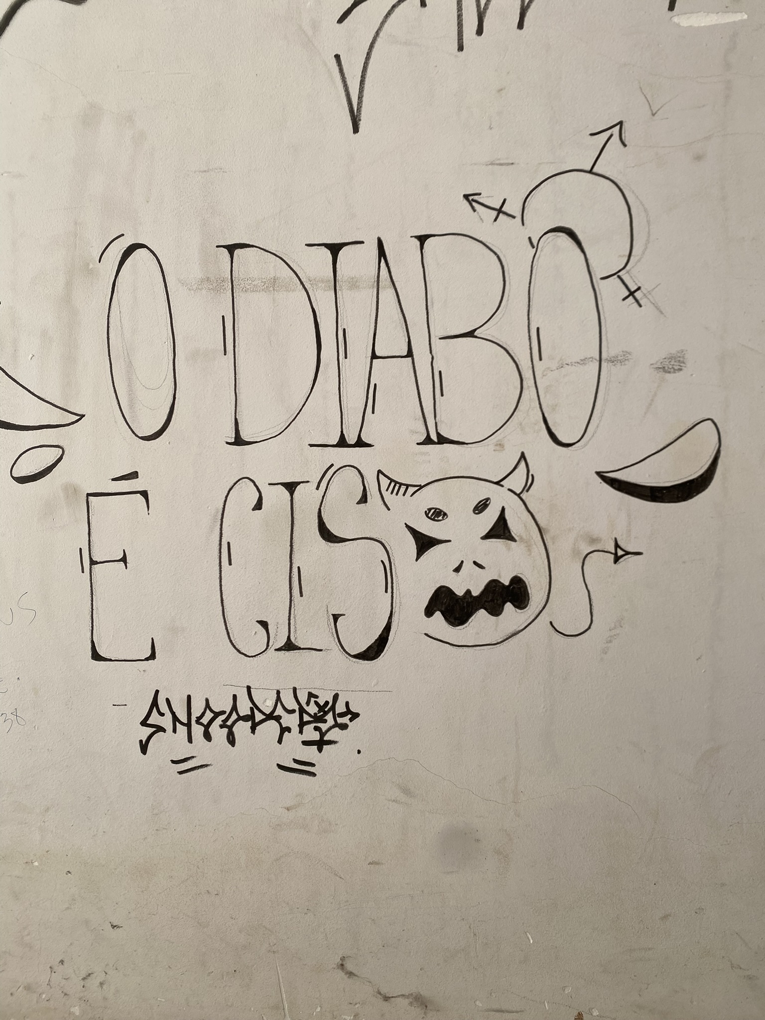 Grafite em uma parede branca com a frase "O DIABO É CIS". As letras são grandes, em contorno preto e estilo de fonte que remete a letras góticas. A palavra "O DIABO" está na linha de cima e "É CIS" na de baixo. Um desenho de uma cabeça de demônio sorridente com chifres, feito em linhas simples, substitui a letra "O" da palavra "CIS".