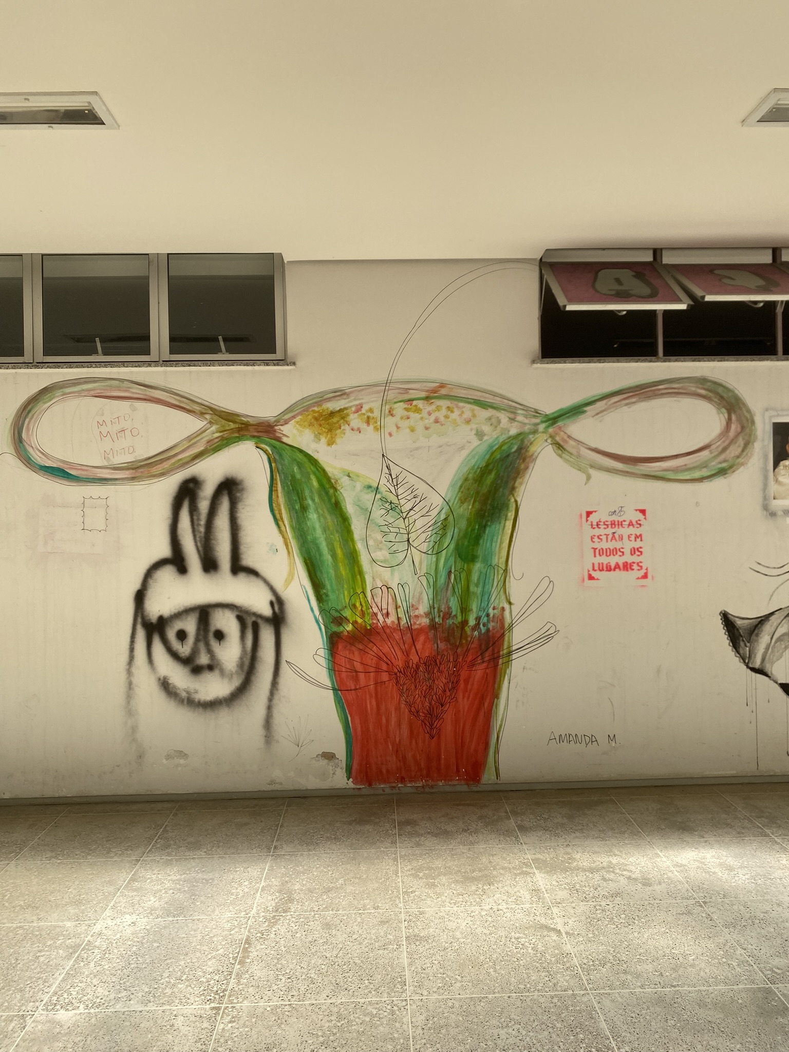 Mural em uma parede branca com janelas na parte superior. O desenho central é uma representação artística dos órgãos reprodutivos femininos, com útero e tubas uterinas estilizados em cores vibrantes, principalmente verde, vermelho e amarelo. O útero está preenchido com um coração vermelho e uma folha.