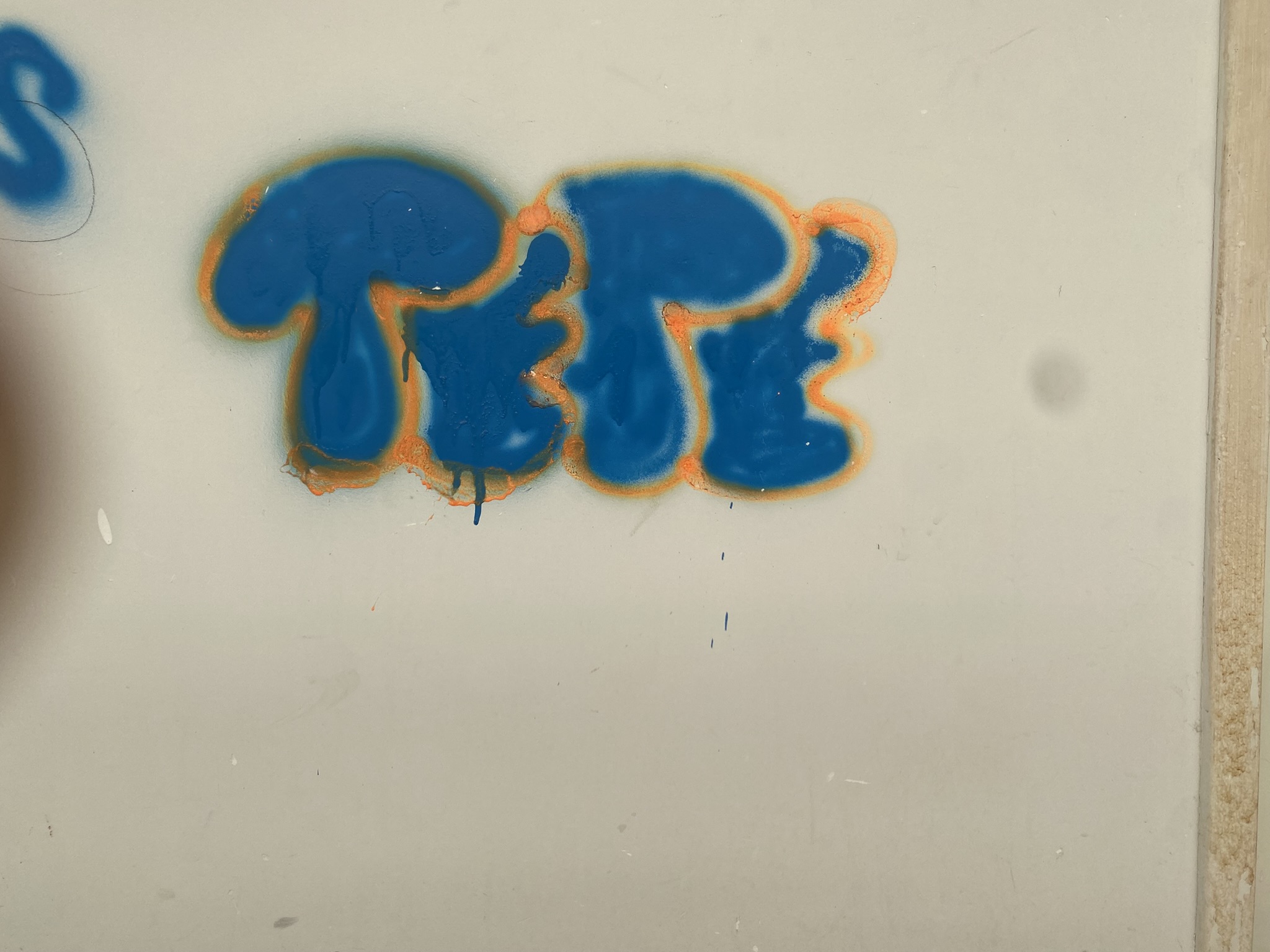 A imagem apresenta uma parede branca com a palavra "TETE" pichada em spray azul vibrante. As letras possuem um contorno esfumaçado em tom alaranjado, criando um efeito de destaque contra o fundo claro. A pichação é centralizada e exibe alguns escorridos sutis de tinta azul na parte inferior.