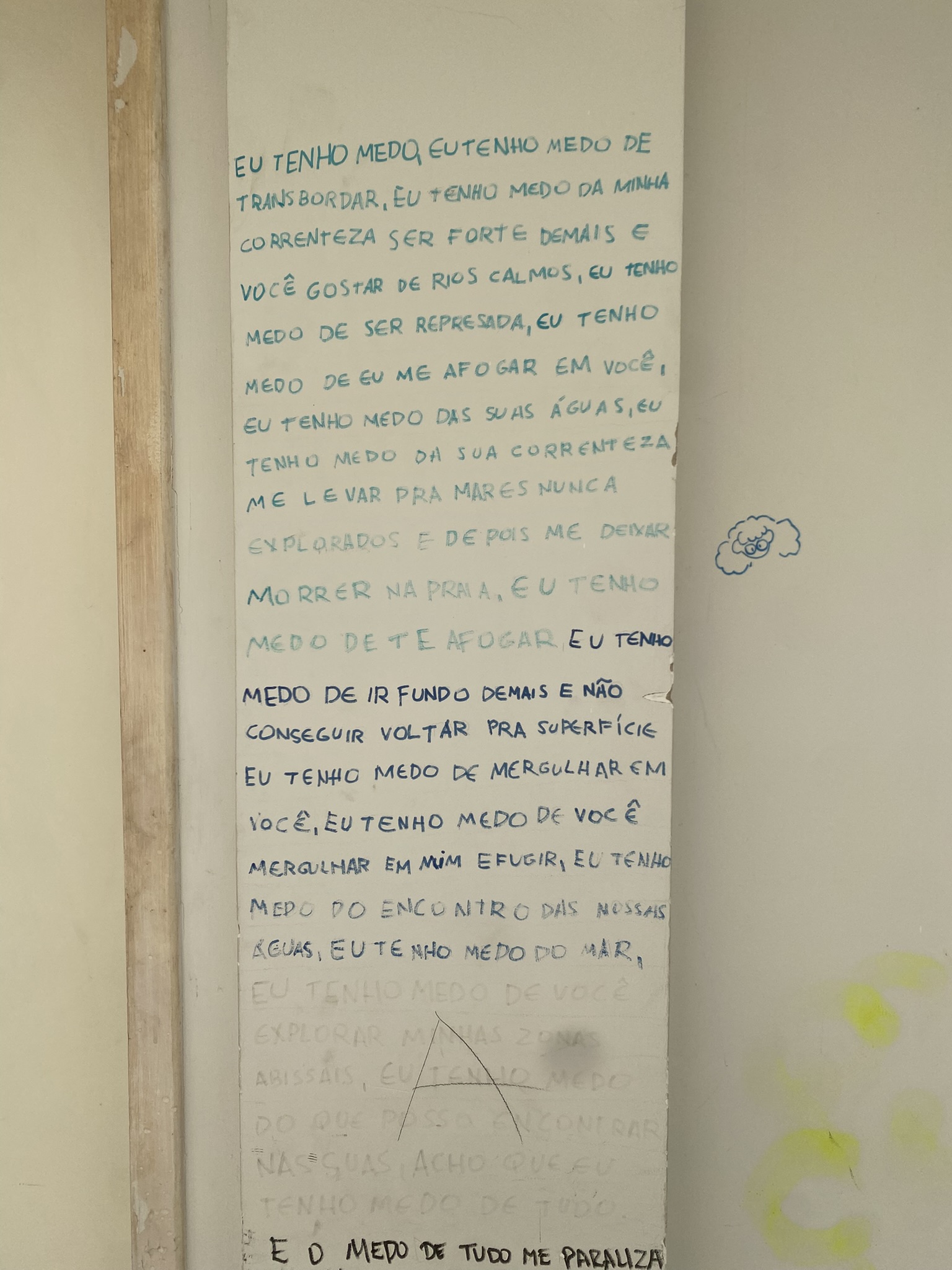 A imagem apresenta um pilar estreito em uma parede clara, onde um longo poema foi escrito à mão com letras maiúsculas em tom azul-esverdeado. O texto utiliza metáforas aquáticas para explorar sentimentos de insegurança e vulnerabilidade em uma relação. O poema diz ""Eu tenho medo, eu tenho medo de transbordar, eu tenho medo da minha correnteza ser forte demais e você gostar de rios calmos, eu tenho medo de ser represada, eu tenho medo de eu me afogar em você, eu tenho medo das suas águas, eu tenho medo da sua correnteza me levar pra mares nunca explorados e depois me deixar morrer na praia. Eu tenho medo de te afogar. Eu tenho medo de ir fundo demais e não conseguir voltar pra superfície. Eu tenho medo de mergulhar em você, eu tenho medo de você mergulhar em mim e fugir, eu tenho medo do encontro das nossas águas, eu tenho medo do mar, eu tenho medo de você explorar minhas zonas abissais, eu tenho medo do que posso encontrar nas suas, acho que eu tenho medo de tudo. E o medo de tudo me paraliza".