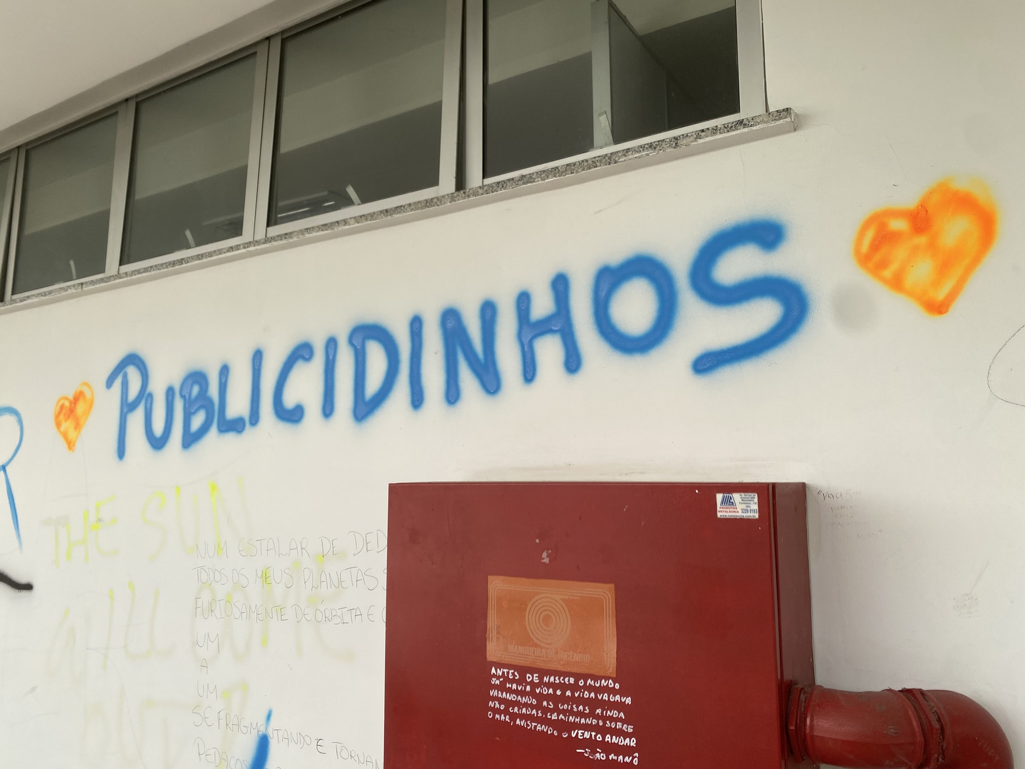 A imagem apresenta uma parede branca com a palavra "PUBLICIDINHOS" pichada em spray azul, acompanhada por dois corações estilizados em tons de amarelo e laranja. Abaixo da pichação, há um armário metálico de hidrante na cor vermelha, que contém um pequeno poema manuscrito em letras brancas.