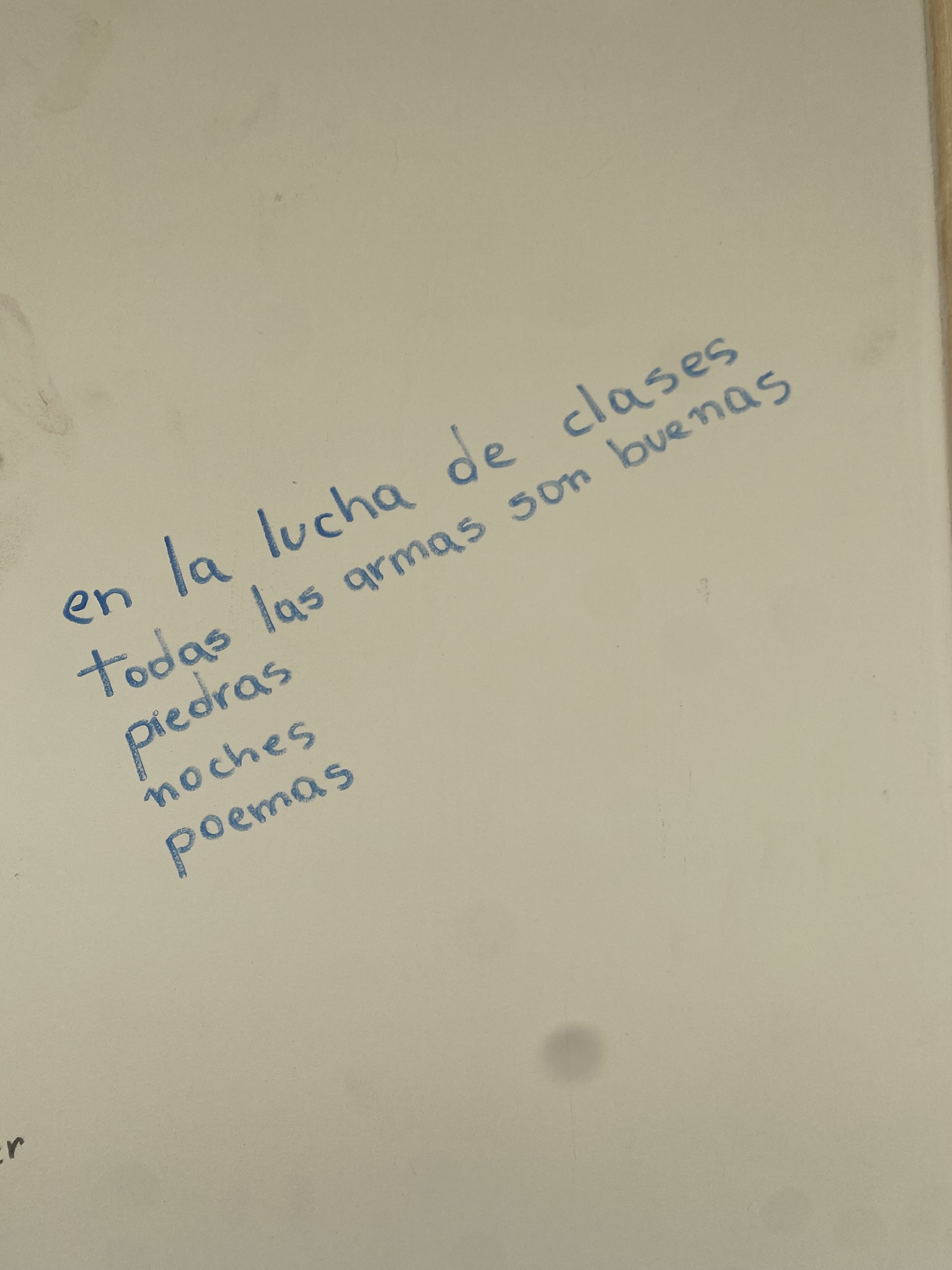 A imagem mostra um pequeno poema escrito à mão com caneta azul sobre uma superfície branca e lisa, que diz: "en la lucha de clases todas las armas son buenas piedras noches poemas".