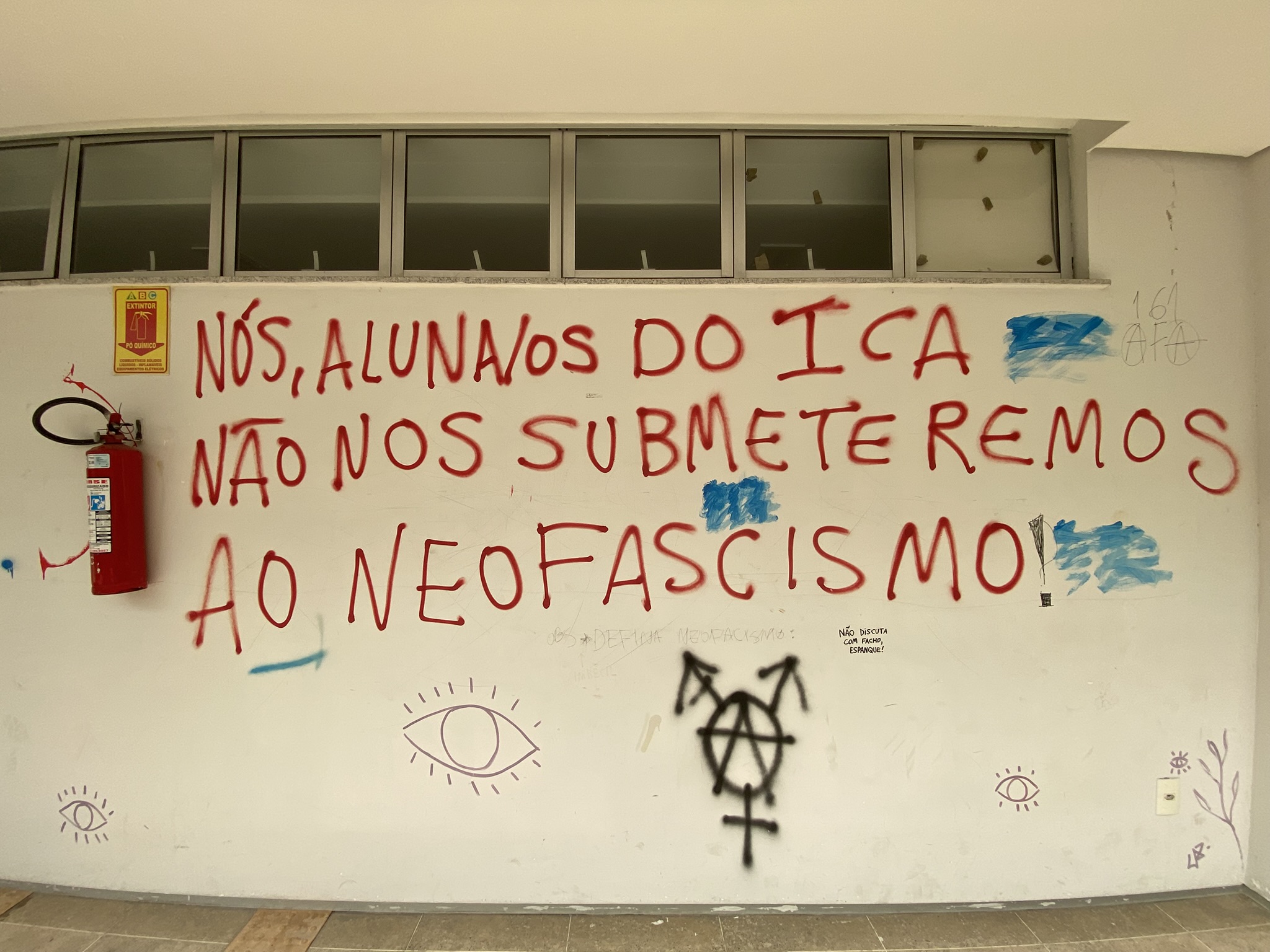 A imagem apresenta uma parede branca com um manifesto político escrito em letras grandes com spray vermelho. O texto diz: "NÓS, ALUNAS/OS DO ICA NÃO NOS SUBMETEREMOS AO NEOFASCISMO!". Abaixo da frase principal, há diversos símbolos e frases menores em preto e azul, incluindo desenhos de olhos, um símbolo de anarquia fundido com setas de gênero.