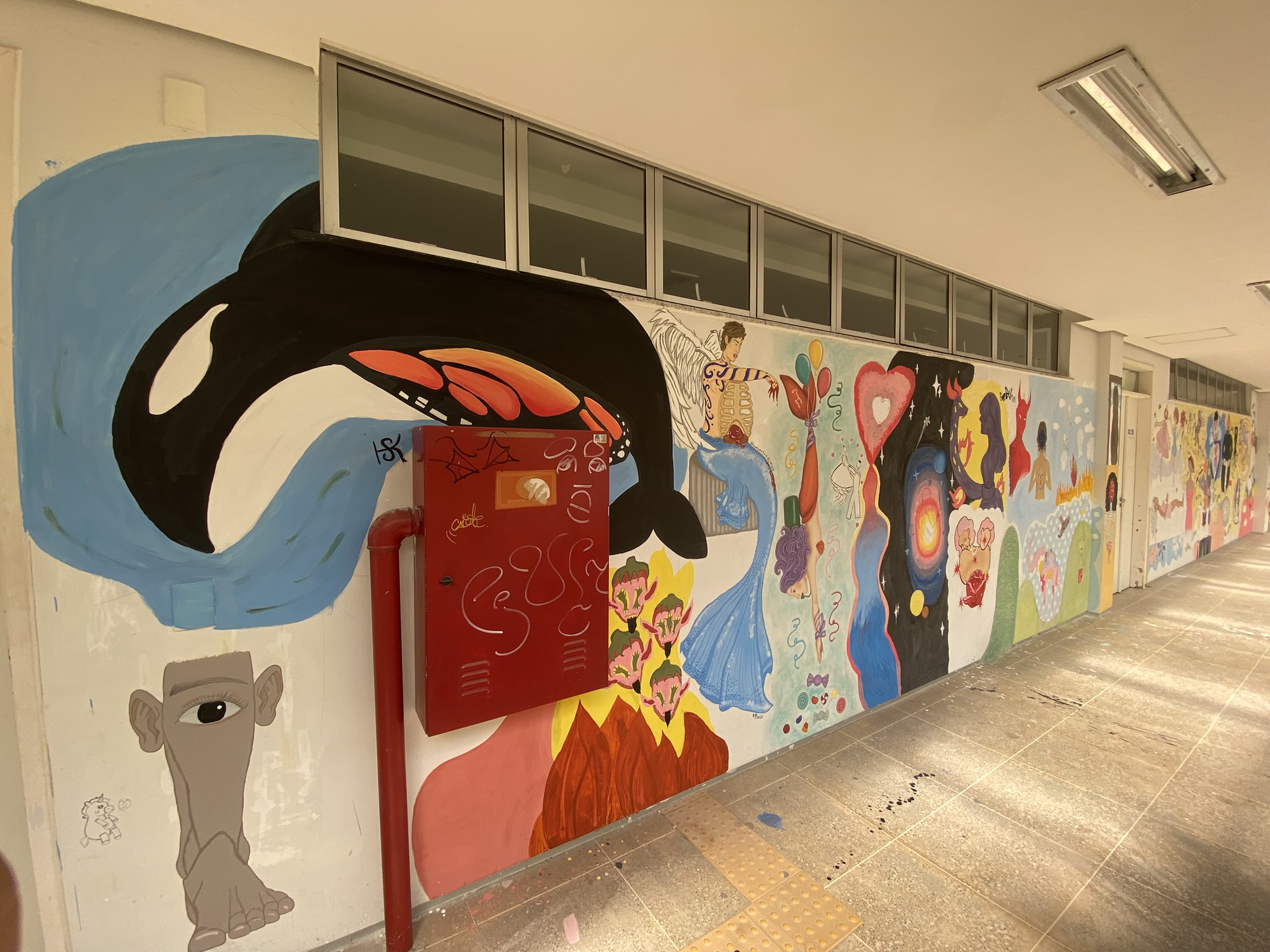 A imagem apresenta um longo corredor do ICA com uma extensa parede branca repleta de murais artísticos coloridos e variados. No primeiro plano, à esquerda, destaca-se uma grande orca preta e branca saltando sobre um fundo azul, próxima a um armário de hidrante vermelho. Abaixo da orca, há o desenho de um pé humano fundido a uma cabeça com um único olho central. O mural continua pelo corredor com diversas figuras, incluindo uma sereia azul, corações, chamas e silhuetas humanas, estendendo-se sob uma fileira de janelas altas.