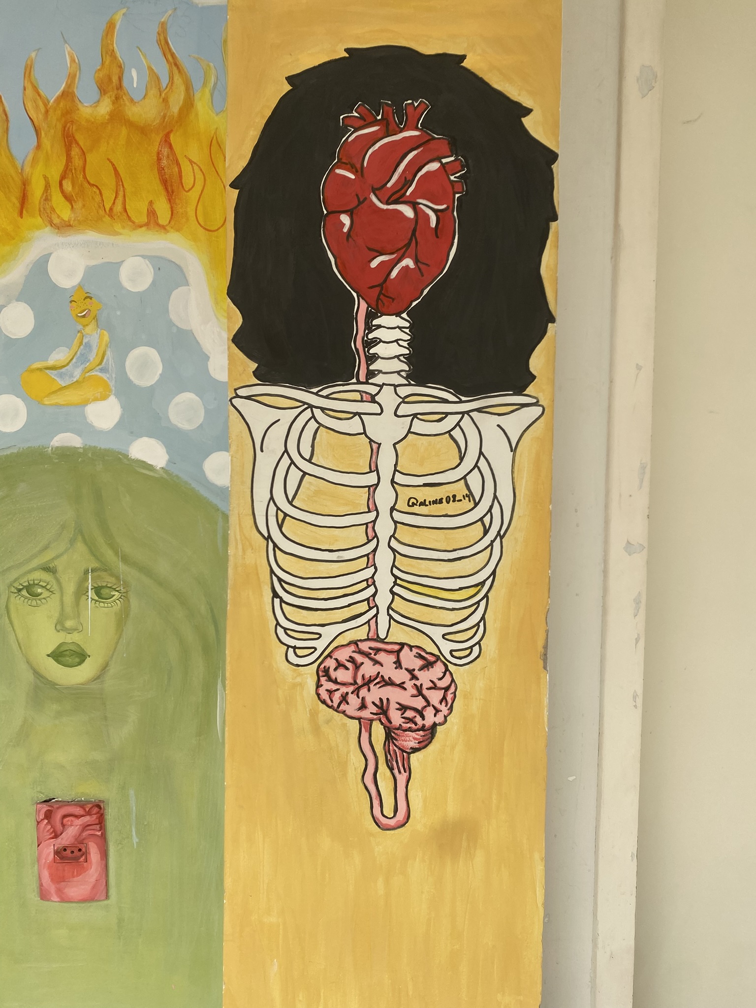 A imagem apresenta uma pintura vertical dividida em duas seções principais sobre uma parede. À direita, sobre um fundo amarelo, observa-se a representação de um esqueleto humano da cintura para cima, encimado por um grande coração anatômico vermelho posicionado onde ficaria a cabeça, contra um fundo de cabelos pretos volumosos. Na base da coluna vertebral, há o desenho de um cérebro rosado. À esquerda, em uma seção de fundo verde e azul, vê-se o rosto de uma mulher de pele esverdeada e longos cabelos claros, acima da qual uma figura amarela pequena medita cercada por chamas e círculos brancos.