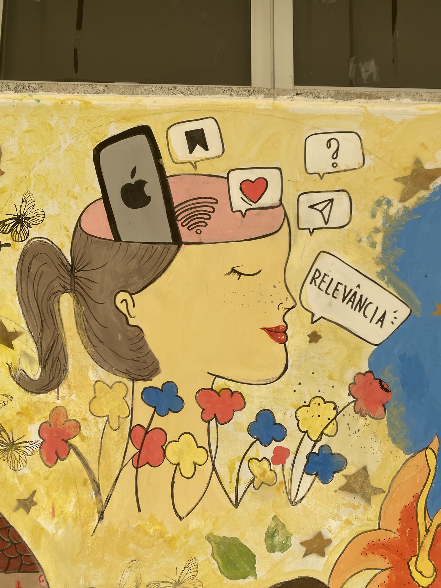 A imagem apresenta a pintura mural de um perfil feminino sobre um fundo amarelo claro salpicado de flores coloridas e estrelas. A cabeça da personagem está aberta no topo, revelando um cérebro rosado de onde emergem diversos ícones digitais, como um smartphone com o logotipo da Apple, um símbolo de Wi-Fi, um balão de coração, um ícone de avião de papel e um ponto de interrogação. À frente do rosto, um balão de fala contém a palavra "RELEVÂNCIA". Borboletas e folhas verdes completam a composição ao redor da figura.