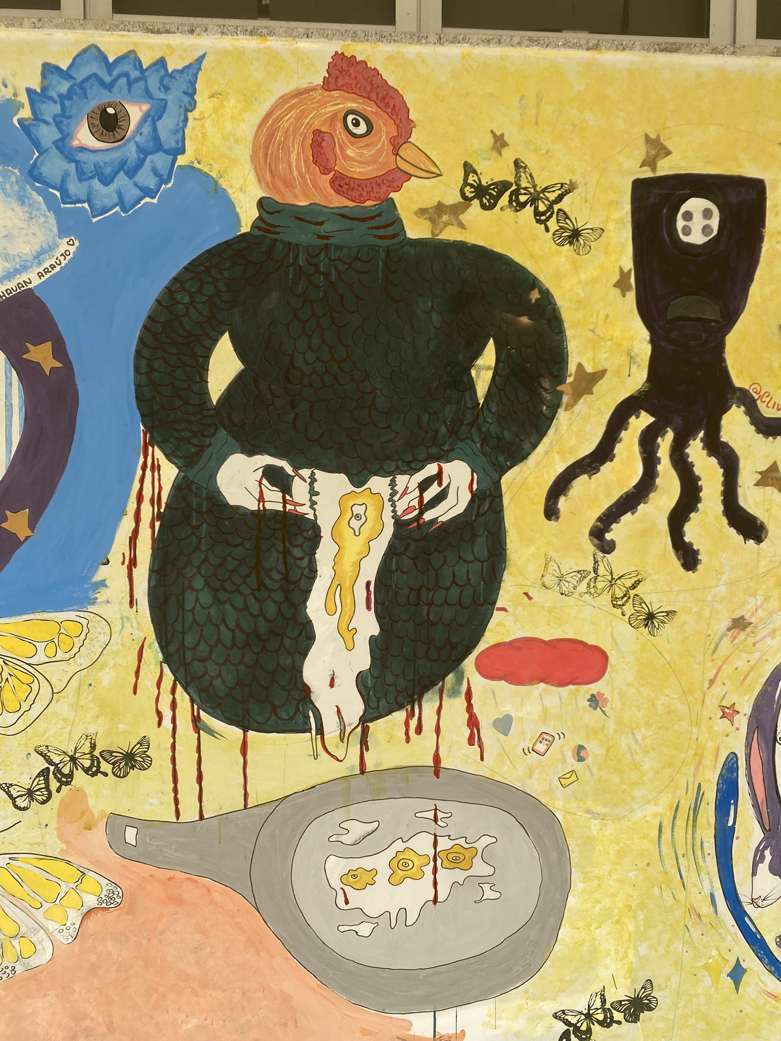 A imagem apresenta uma pintura mural surrealista sobre um fundo amarelo vibrante e texturizado. No centro, destaca-se uma figura corpulenta vestindo uma roupa escura com textura de escamas, possuindo uma cabeça de galinha ruiva. A figura segura uma grande casca de ovo branca da qual escorre um líquido amarelo e sangue, que cai em uma frigideira cinza no chão contendo três ovos fritos com olhos no lugar das gemas. Ao redor, há diversos elementos flutuantes, como borboletas pretas e amarelas, estrelas douradas, uma nuvem vermelha, ícones de redes sociais e uma criatura negra com tentáculos e um único olho que se assemelha a um botão.