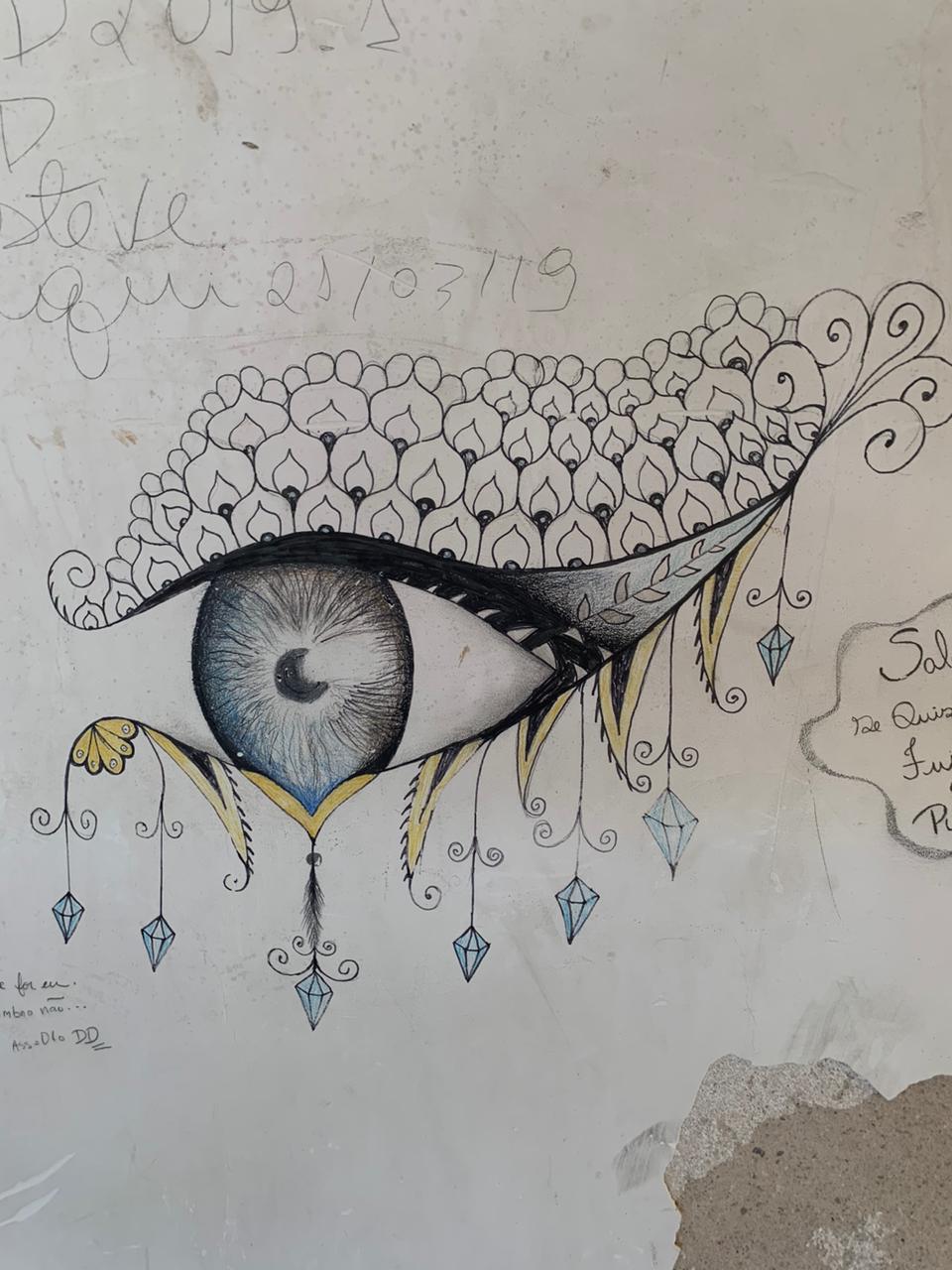 A imagem apresenta um desenho detalhado de um olho humano feito com grafite e lápis de cor sobre uma superfície clara. A parte superior do olho é adornada por uma estrutura ornamental que lembra escamas de peixe ou penas, com padrões repetitivos e espirais. Da parte inferior, pendem diversos pingentes em formato de cristais azuis e elementos decorativos em curvas.