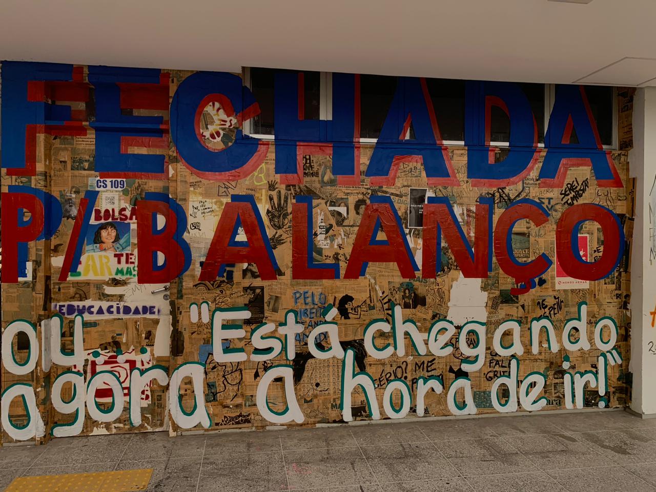 A imagem apresenta uma porta e parte de uma parede inteiramente cobertas por uma colagem de jornais e revistas. Sobre essa textura, destaca-se a frase "FECHADA P/ BALANÇO" escrita em letras garrafais, alternando entre azul e vermelho. Na parte inferior, uma segunda frase em letras brancas com contorno preto diz: "ou agora 'Está chegando a hora de ir!'".