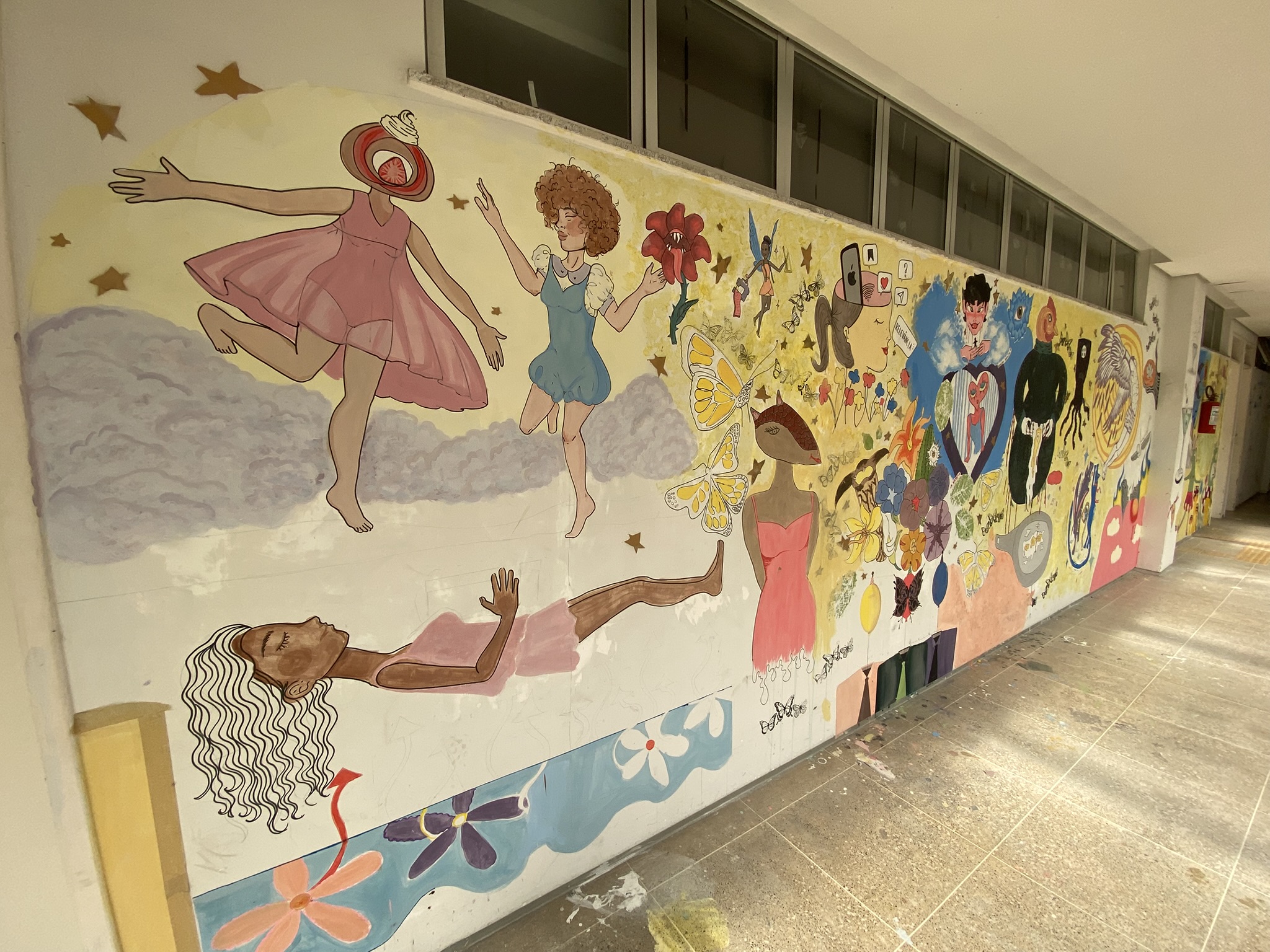 A imagem apresenta um corredor do ICA com uma parede branca repleta de murais artísticos contínuos. As pinturas mostram diversas figuras femininas em situações surreais: uma mulher com cabeça de doce flutuando sobre nuvens, outra figura correndo e uma terceira deitada na parte inferior. A composição segue com diversos elementos menores, como borboletas, flores, corações e ícones digitais, estendendo-se por toda a extensão da parede sob as janelas.