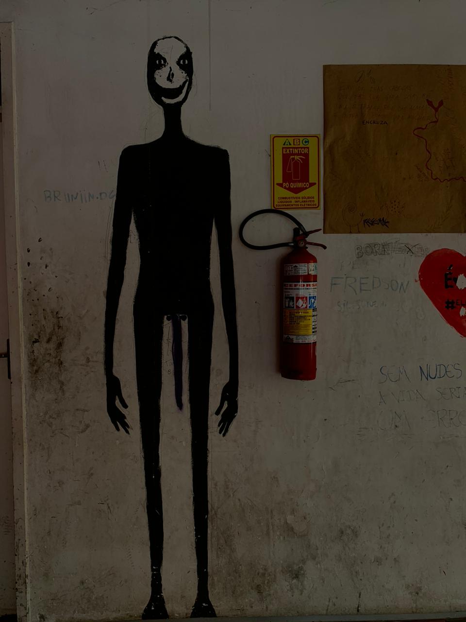 A imagem apresenta uma parede branca com diversas inscrições e a silhueta de uma figura alta e esguia pintada inteiramente de preto. A figura possui um rosto branco com um sorriso largo e olhos vazios, e da região da pélvis pende um elemento longo e fino em tom roxo.
