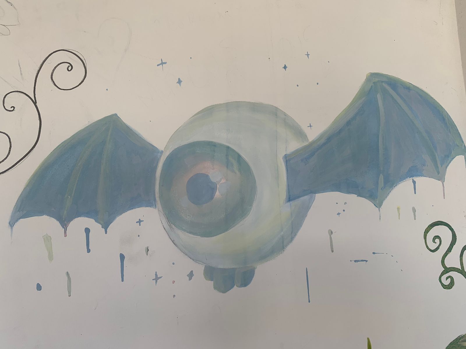 A imagem apresenta, sobre um fundo branco, a pintura de um grande globo ocular alado. O olho possui uma íris em tons de azul e cinza, e dele partem duas grandes asas de morcego em azul-claro com detalhes sombreados. Pequenas estrelas de quatro pontas e gotas de tinta azul e cinza estão espalhadas ao redor da figura, criando um efeito de flutuação.