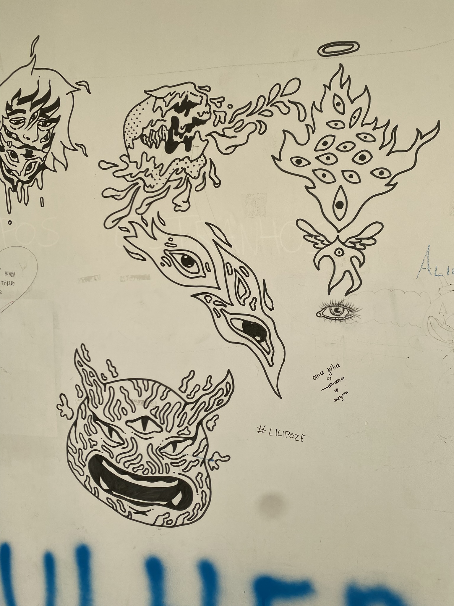 A imagem apresenta uma parede branca com diversos desenhos feitos em traço preto, lembrando o estilo de tatuagens flash. Destacam-se uma cabeça de demônio com três olhos e padrões ondulados, um coração formado por vários olhos, chifres e chamas, e uma figura angelical minimalista com auréola e asas.