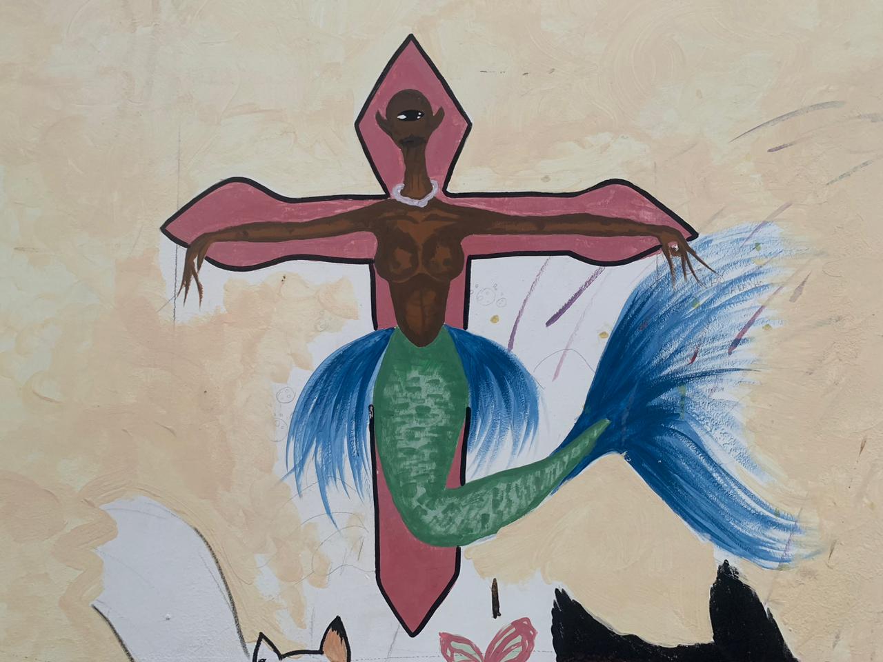 A imagem apresenta uma pintura mural de uma sereia negra crucificada em uma cruz rosa sobre um fundo bege texturizado. A sereia possui um único olho centralizado na testa, braços abertos em posição de crucificação e uma cauda verde que termina em uma grande nadadeira azul vibrante.