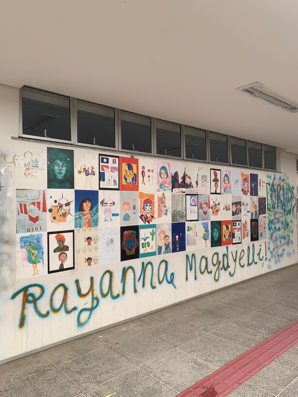 A imagem apresenta um corredor institucional com uma parede branca decorada por uma grade de diversos cartazes e ilustrações artísticas coloridas. Abaixo dos cartazes, o nome "Rayanna Magdyelli!"