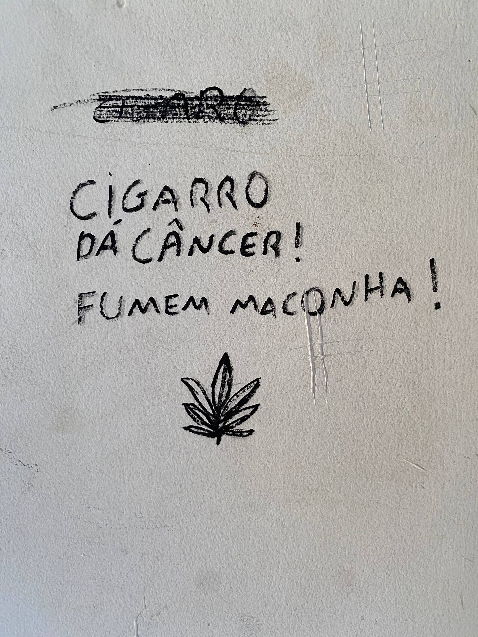A imagem apresenta uma mensagem manuscrita em letras maiúsculas com marcador preto sobre uma parede branca. O texto afirma: "CIGARRO DÁ CÂNCER! FUMEM MACONHA!". Logo abaixo das frases, há o desenho detalhado de uma folha de cannabis com sete pontas.