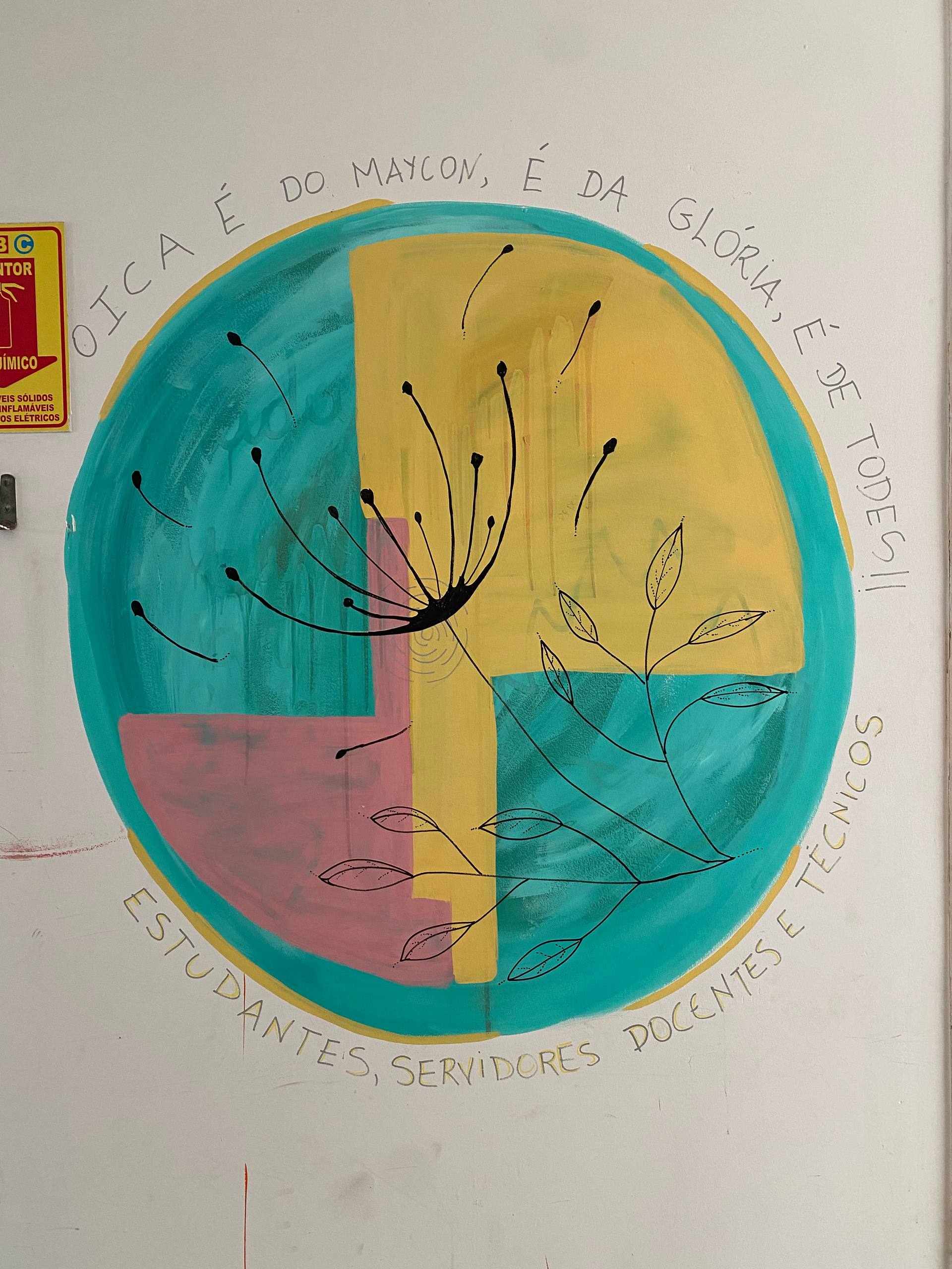 A imagem apresenta um mural circular colorido pintado sobre uma parede branca. O círculo é dividido em quatro quadrantes com cores de fundo diferentes: verde-água, amarelo, rosa e azul-petróleo. Sobre esse fundo, há desenhos em traços pretos finos de dentes-de-leão cujas sementes parecem flutuar e ramos com folhas delicadas. Ao redor da borda superior do círculo, está escrita a frase manuscrita: "O ICA É DO MAYCON, É DA GLÓRIA, É DE TODES!!". Na borda inferior, lê-se: "ESTUDANTES, SERVIDORES DOCENTES E TÉCNICOS".