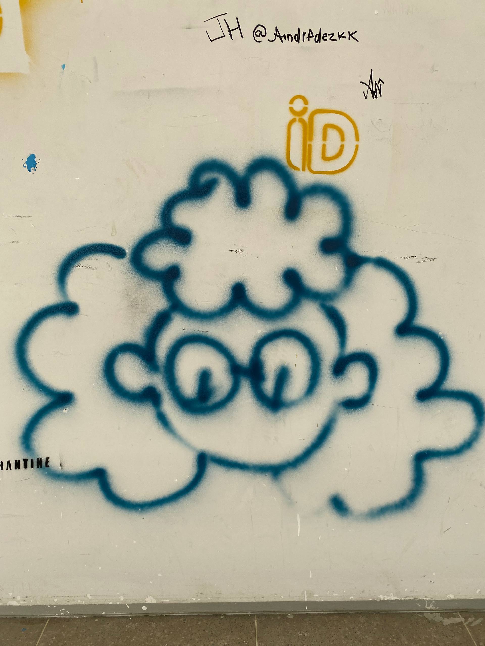 A imagem apresenta um desenho estilizado pichado com spray azul sobre uma parede branca. A figura retrata o rosto de um personagem com óculos redondos e cabelos volumosos e cacheados, representados por contornos arredondados.