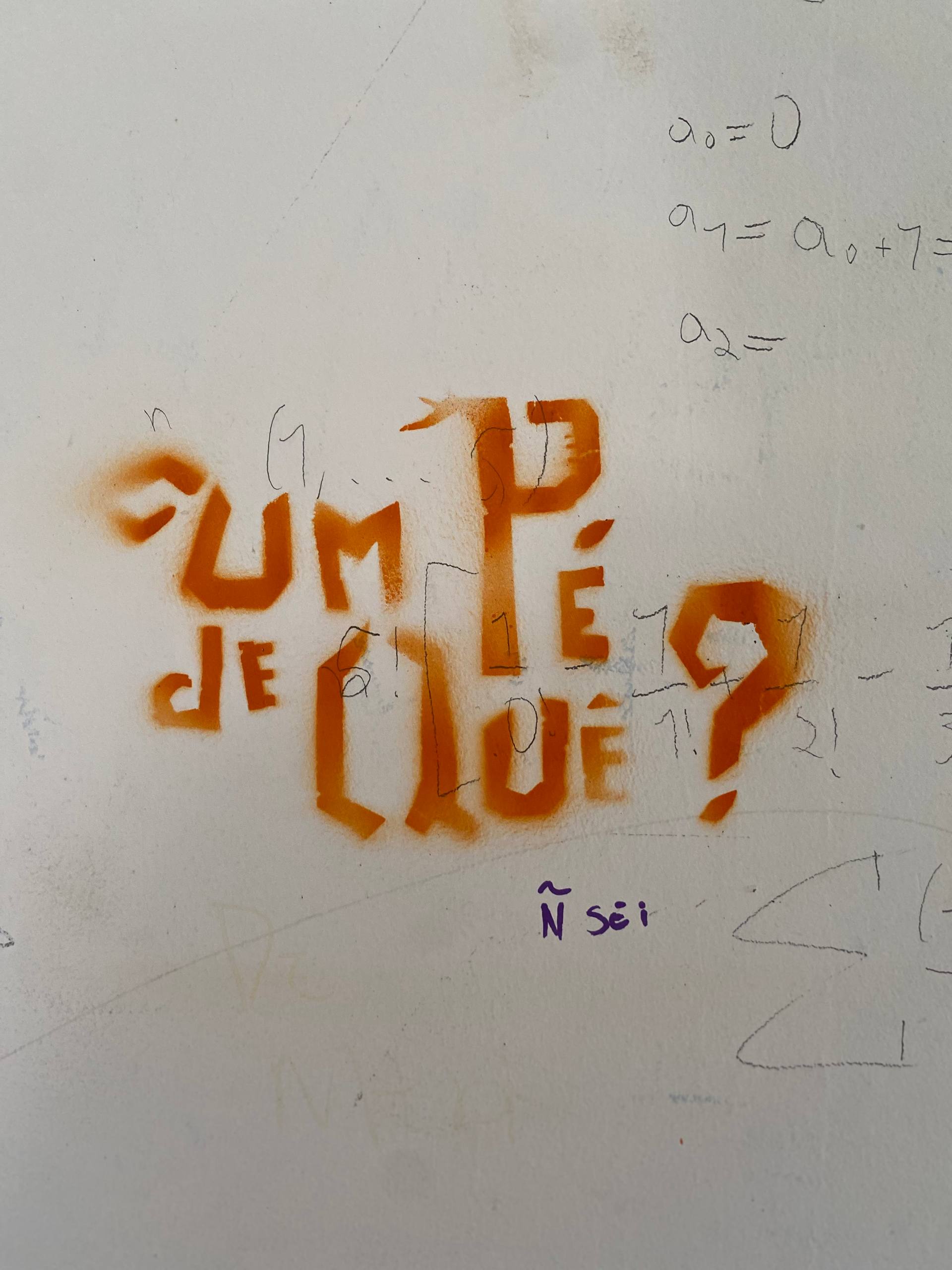 A imagem apresenta uma pichação feita em spray laranja sobre uma parede branca com diversas anotações matemáticas a lápis ao fundo. O texto, escrito em letras estilizadas e fragmentadas, pergunta: "CUM PÉ DE QUÊ?". Logo abaixo da interrogação, alguém acrescentou de forma manuscrita com caneta roxa a resposta: "Ñ sei".
