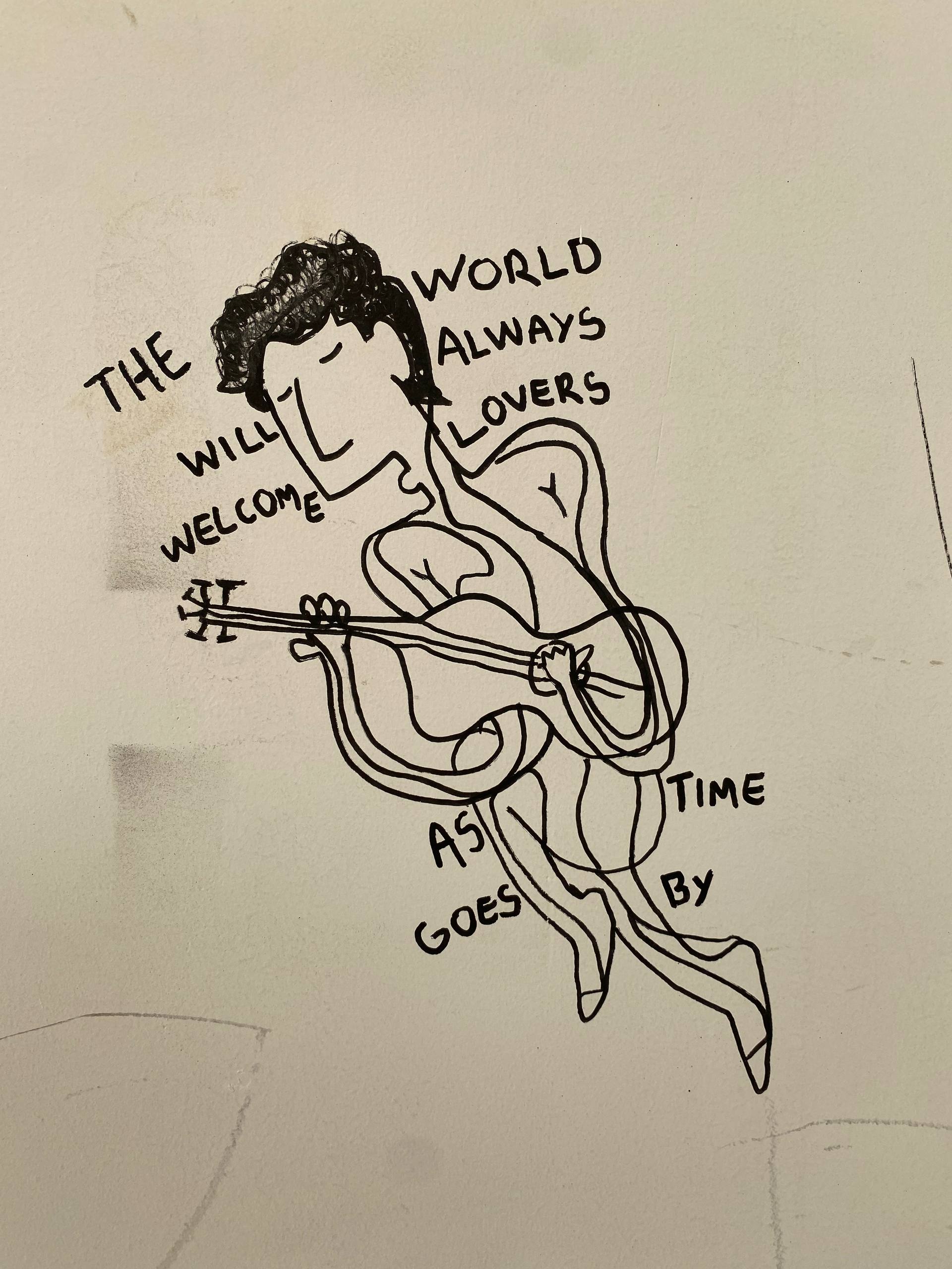 A imagem apresenta um desenho linear feito com marcador preto sobre uma parede branca. A ilustração retrata uma figura humana estilizada com corpo fluido e ondulado, segurando e tocando o que parece ser um violão ou um instrumento de cordas. Integradas ao desenho e contornando a figura, estão as palavras em inglês: "THE WORLD WILL ALWAYS WELCOME LOVERS AS TIME GOES BY" (O mundo sempre acolherá os amantes conforme o tempo passa).