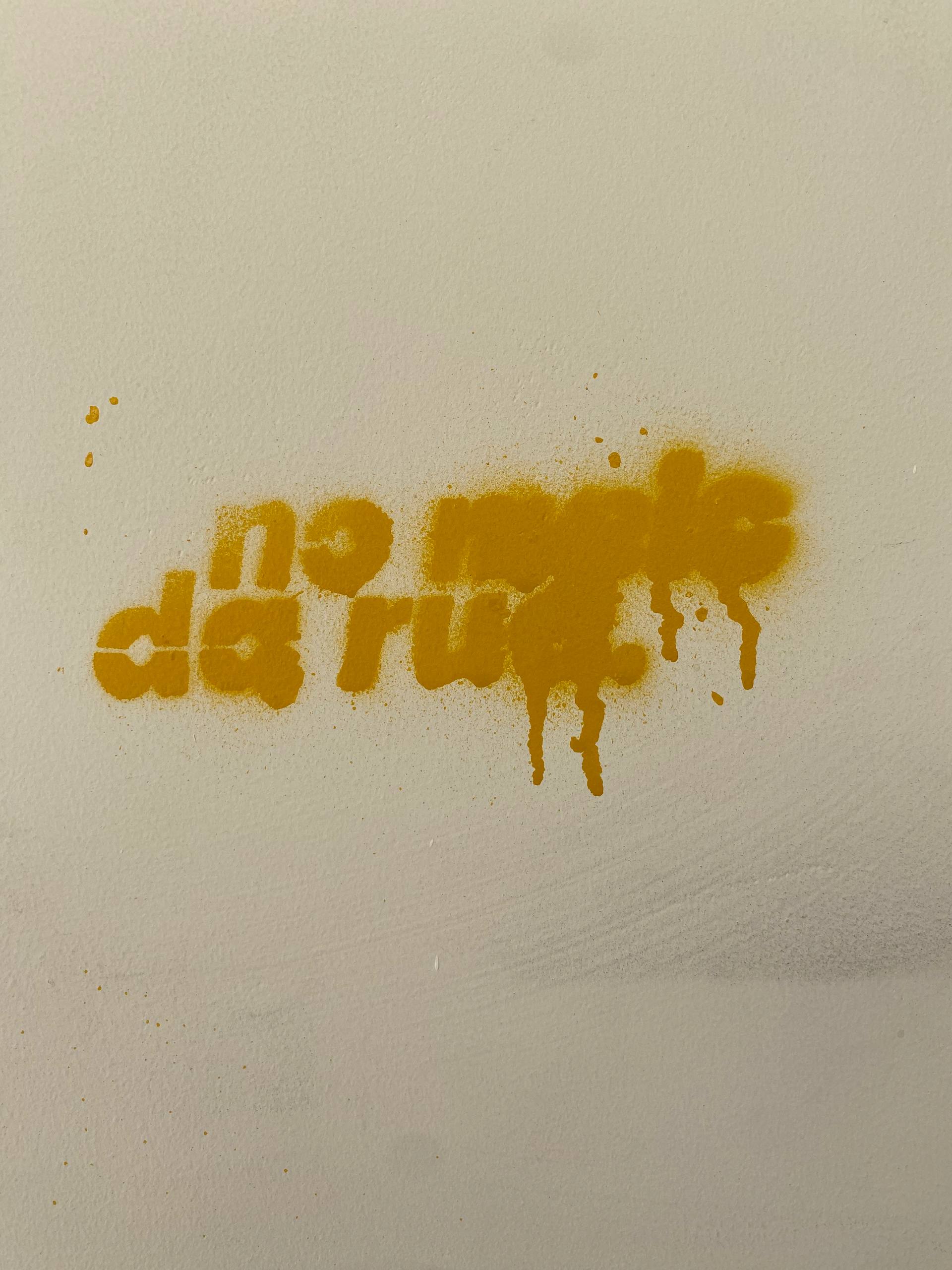 A imagem apresenta uma pichação feita com a técnica de estêncil em spray amarelo vibrante sobre uma parede clara. O texto, escrito em letras arredondadas e com alguns borrões de tinta que escorrem para baixo, diz: "no rastro da rua". A aplicação da cor amarela cria um contraste suave com o fundo branco da parede, que possui uma textura levemente granulada.