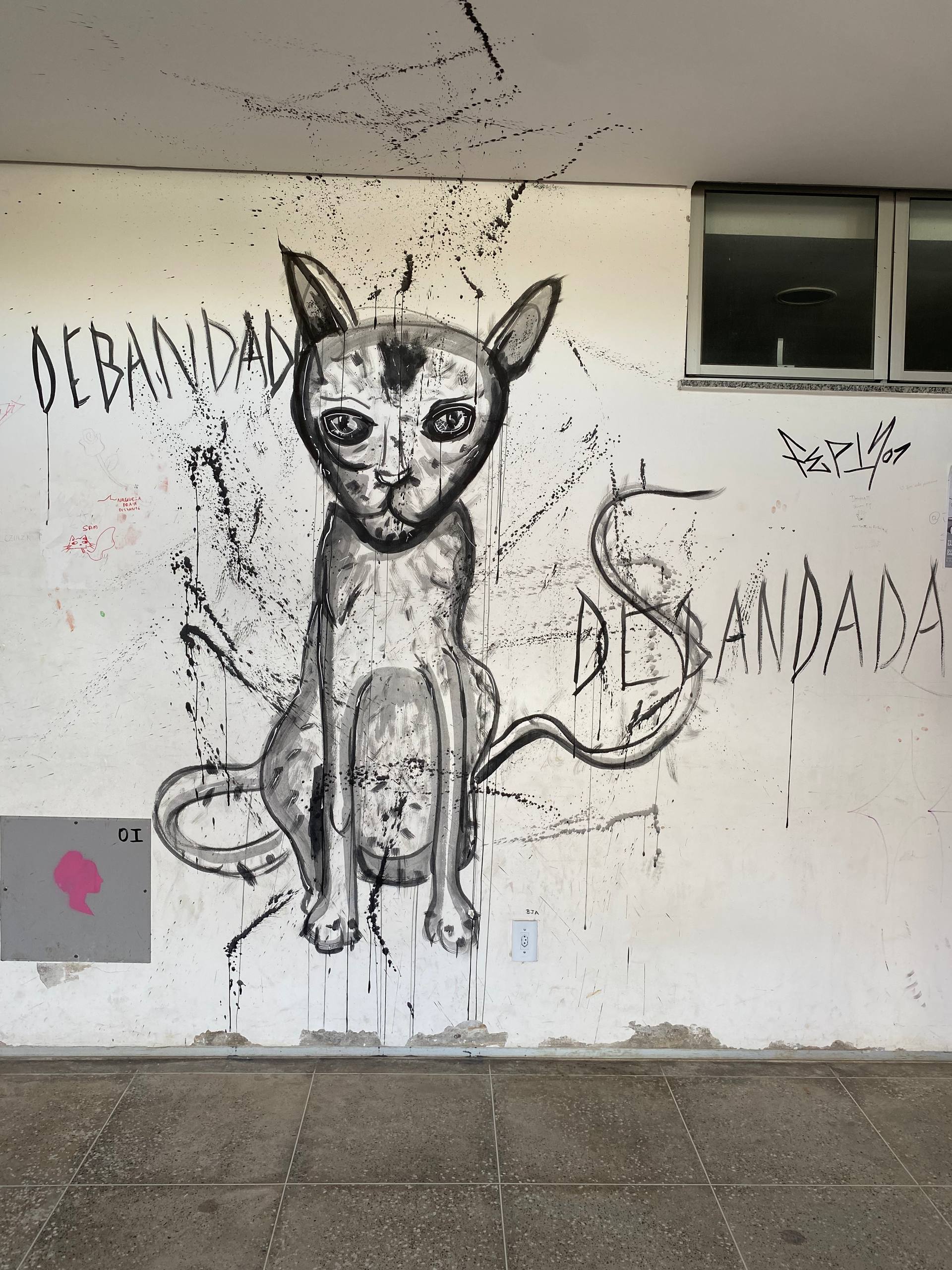 A imagem apresenta um grande grafite feito com traços pretos expressivos sobre uma parede branca. O desenho central retrata um gato magro, com olhos grandes e marcantes, sentado em uma postura frontal. Ao redor do animal, a palavra "DEBANDADA" está escrita em letras grandes e angulares em ambos os lados. A obra exibe um estilo artístico cru, com pingos e respingos de tinta preta que escorrem pela parede, conferindo uma sensação de movimento.