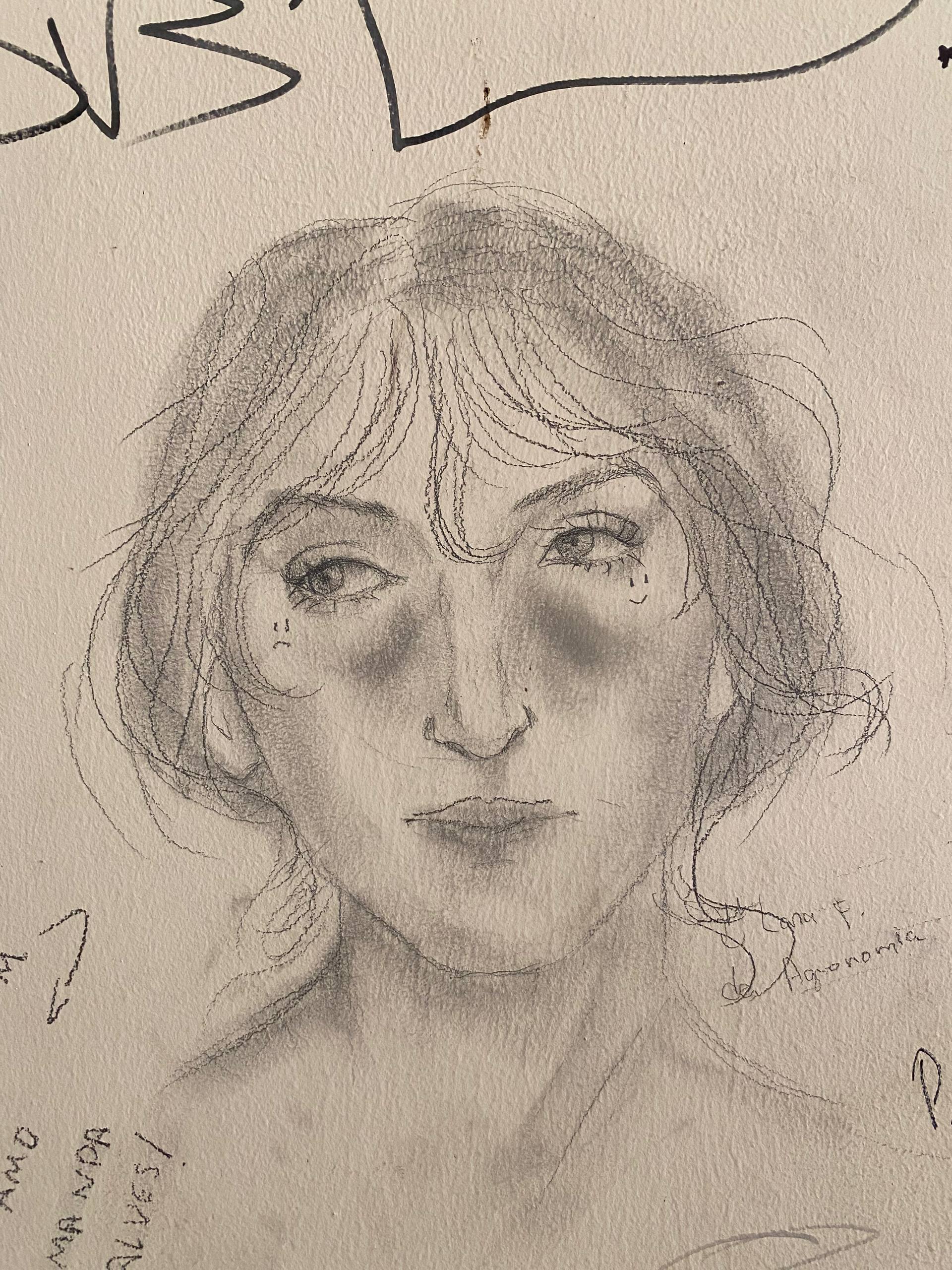 A imagem apresenta o retrato de uma pessoa desenhado a lápis sobre uma superfície clara. O desenho mostra feições delicadas, com olhos grandes voltados para o lado e cabelos ondulados. Nas bochechas, há pequenas marcas decorativas: dois traços que lembram uma expressão triste abaixo do olho esquerdo e uma pequena carinha sorridente abaixo do olho direito.