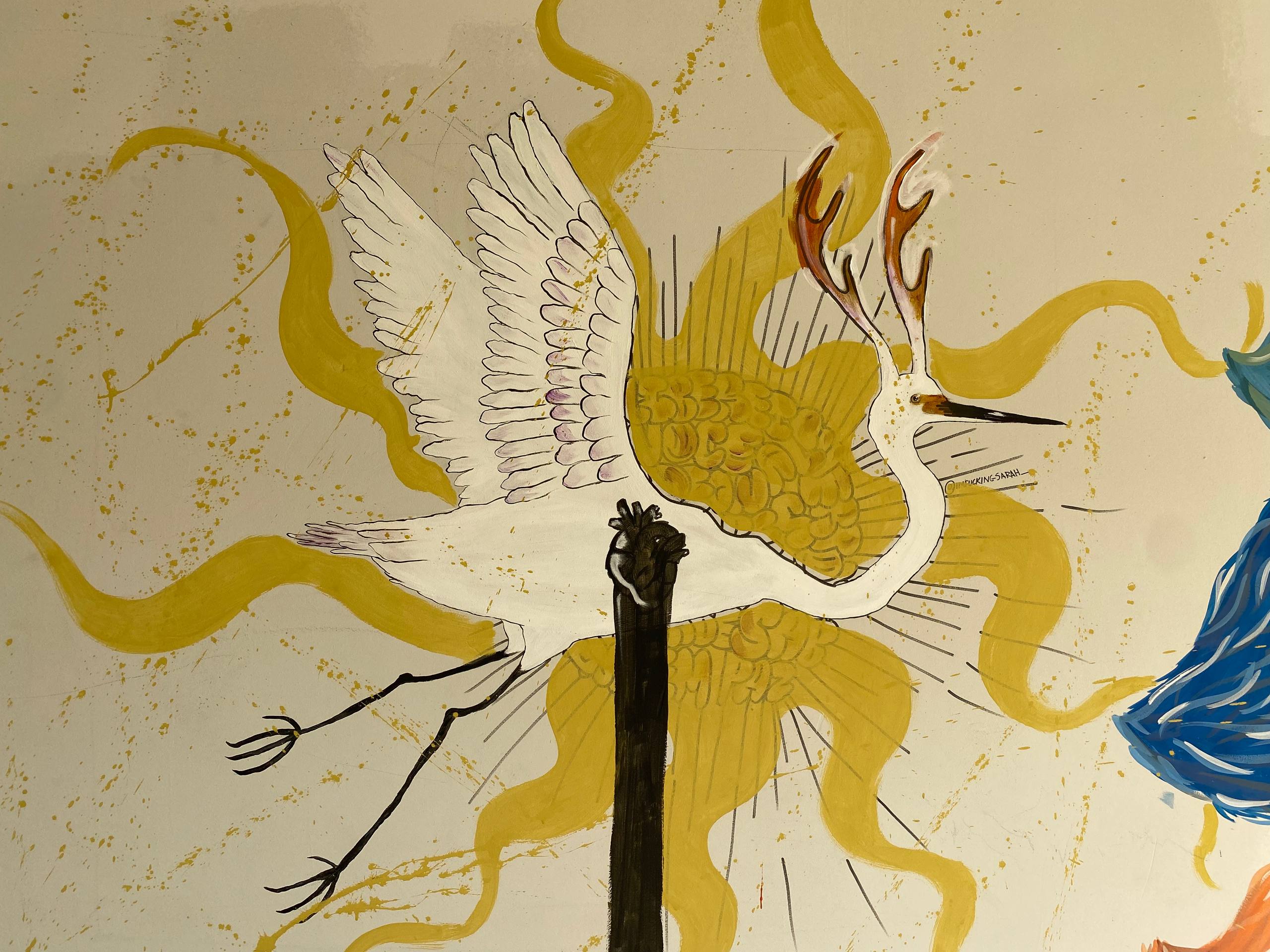 A imagem apresenta uma pintura mural de uma garça branca estilizada em pleno voo. A ave possui chifres de cervo ramificados no topo da cabeça e é atravessada verticalmente por um poste ou tronco preto que possui um coração anatômico no topo. Ao fundo, formas onduladas e explosivas em amarelo sugerem a presença de um sol ou luz intensa.