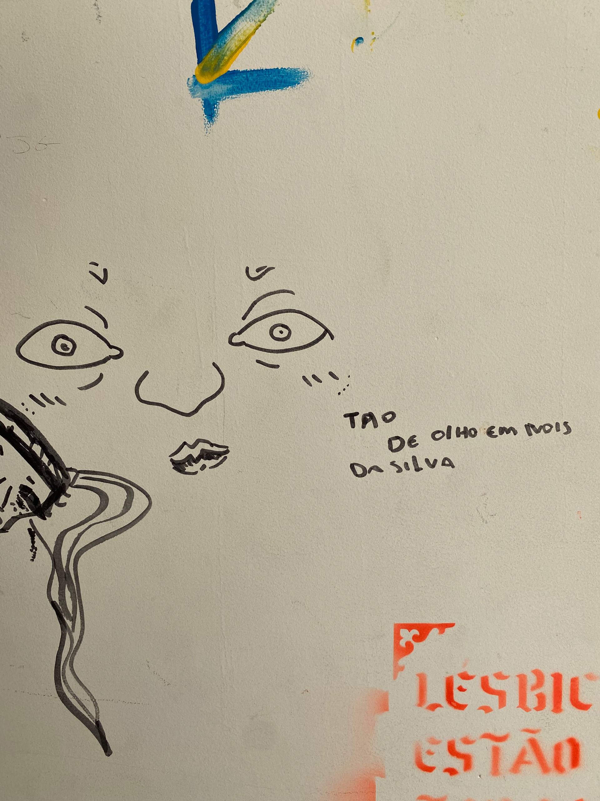 A imagem apresenta esboços e inscrições em uma parede branca. Centralizado, há o desenho minimalista de um rosto humano composto apenas por olhos arregalados, sobrancelhas arqueadas, um contorno simples de nariz e lábios pequenos.