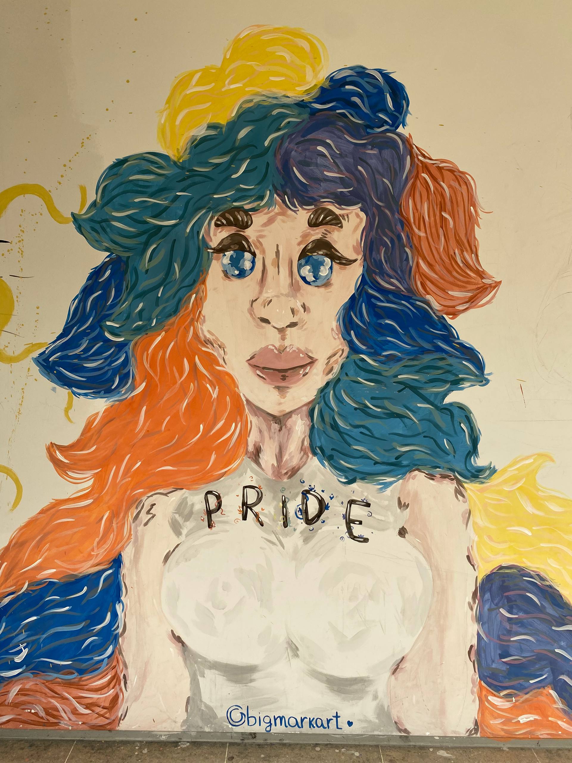 A imagem apresenta uma pintura mural colorida de um busto feminino sobre uma parede branca. A personagem possui pele clara e cabelos volumosos pintados com faixas horizontais em azul, amarelo e laranja. Seus olhos são grandes, azuis e brilhantes, com cílios marcados. No peito da figura, está escrita a palavra "PRIDE" em letras pretas maiúsculas. Na base da pintura, nota-se a assinatura "@bigmarkart".