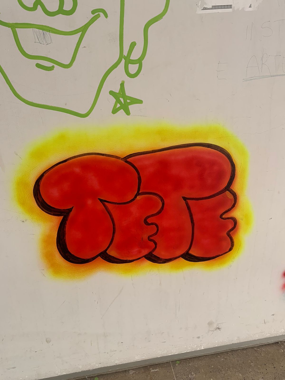 Esta é uma imagem em close-up de uma parte de uma parede branca onde há um grafite estilo bubble letter em destaque. O grafite principal é uma palavra curta ou um conjunto de letras que formam a palavra TETE em vermelho vibrante com contorno preto e um efeito tridimensional arredondado. Este throw-up está sobreposto a um outline em spray que irradia em tons de amarelo e laranja, criando um brilho em torno das letras.