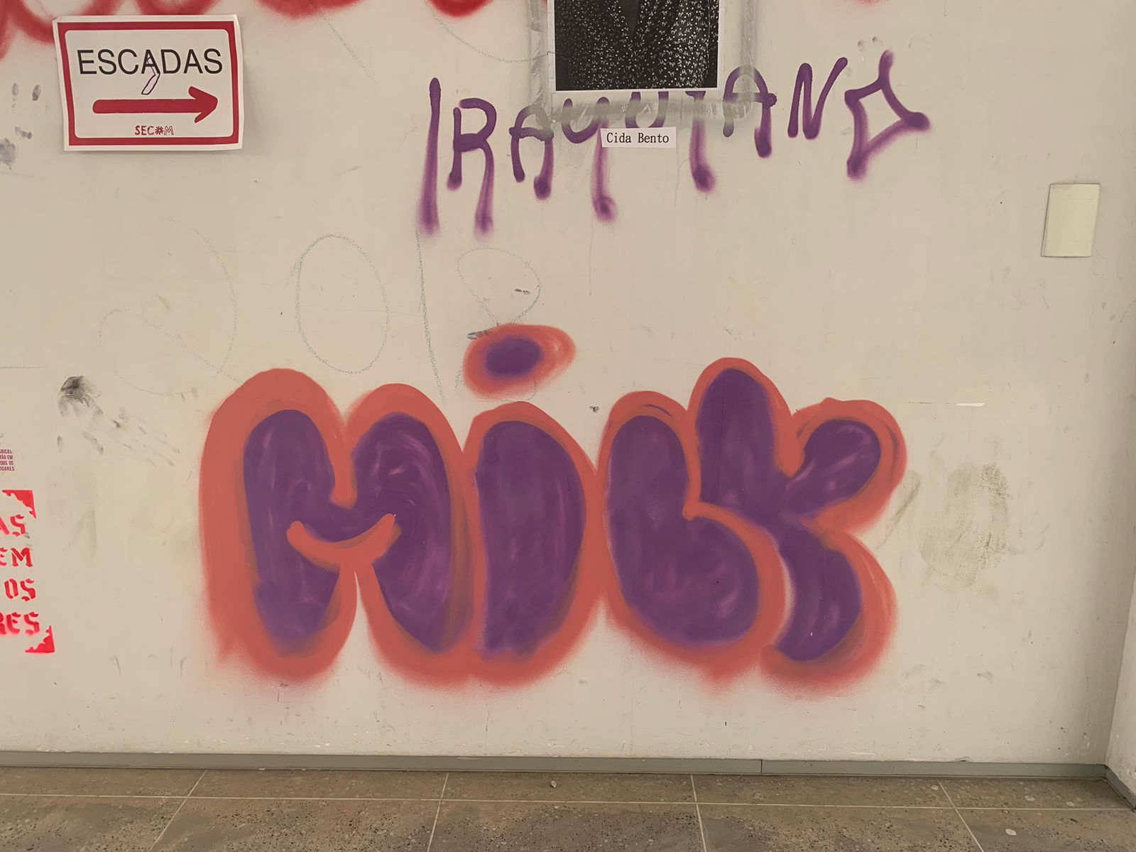 Na foto em close-up, uma parede branca é o suporte para várias intervenções urbanas. Em destaque, um throw-up em letras bolha (bubble style) forma a palavra "MILK" em spray. As letras são preenchidas com tinta roxa e contornadas com uma camada de spray laranja-avermelhado.