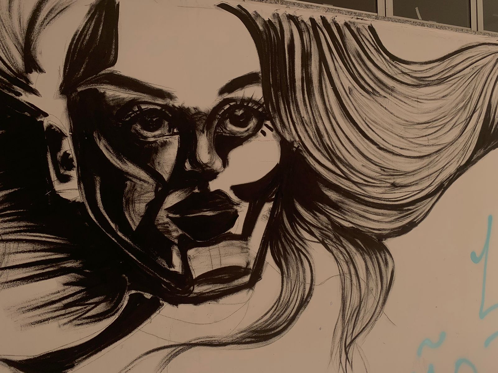 Esta é uma foto em close-up de uma arte em preto e branco, que ocupa a maior parte da imagem. O desenho é um retrato estilizado e expressivo de um rosto feminino, feito com traços fortes e dinâmicos, que criam sombras acentuadas e formas geométricas no rosto para dar profundidade e drama. O cabelo da figura é representado com longas linhas onduladas que parecem esvoaçar para o lado direito.