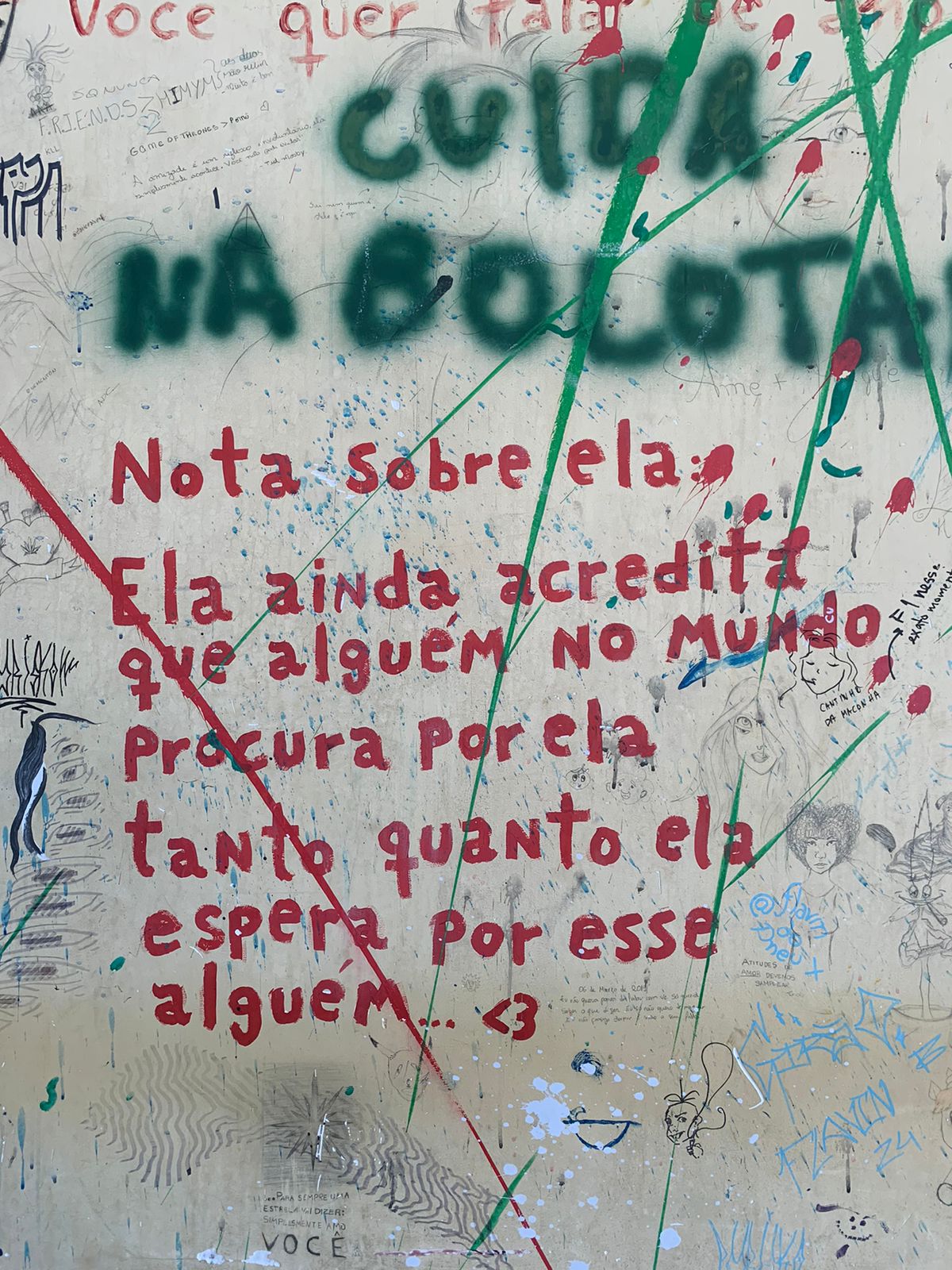 Uma parede amarelada, intensamente coberta por grafites e marcas de tinta, apresenta várias camadas de intervenções. O centro da imagem é dominado por um poema em spray vermelho que diz: "Nota sobre ela: Ela ainda acredita que alguém no mundo procura por ela tanto quanto ela espera por esse alguém... <3". Ao redor, a parede está repleta de outras escritas em várias cores e estilos.