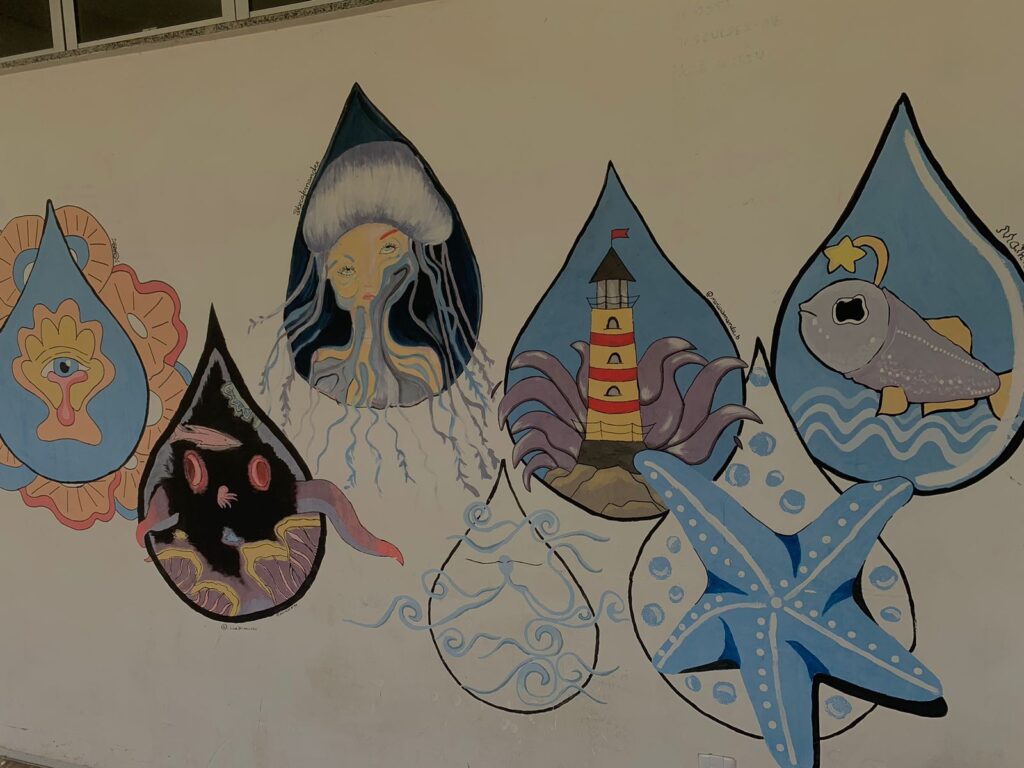 Mural artístico em uma parede branca, apresentando uma composição de gotas d'água estilizadas, cada uma contendo ilustrações com temas marinhos e fantásticos. Da esquerda para a direita: a primeira gota é azul-claro com contorno laranja e contém um olho estilizado com asas; a segunda é escura, quase preta, e mostra criaturas marinhas e algas vermelhas; a gota central é escura no topo e retrata o busto de uma mulher-água-viva com cabelos fluidos em tons de amarelo e cinza; a gota seguinte, com fundo azul-claro, contém um farol listrado de vermelho e branco sobre rochas com tentáculos roxos ao redor; a gota mais à direita mostra um peixe cinza-azulado com uma estrela na cabeça, sobre ondas. Entre e sob as gotas, há elementos adicionais como uma estrela-do-mar azul com bolhas e contornos de ondas.