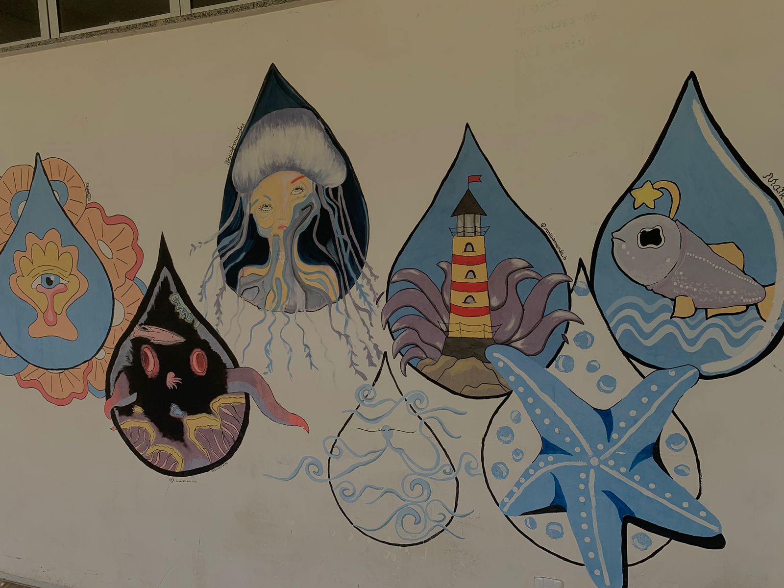 Mural artístico em uma parede branca, apresentando uma composição de gotas d'água estilizadas, cada uma contendo ilustrações com temas marinhos e fantásticos. Da esquerda para a direita: a primeira gota é azul-claro com contorno laranja e contém um olho estilizado com asas; a segunda é escura, quase preta, e mostra criaturas marinhas e algas vermelhas; a gota central é escura no topo e retrata o busto de uma mulher-água-viva com cabelos fluidos em tons de amarelo e cinza; a gota seguinte, com fundo azul-claro, contém um farol listrado de vermelho e branco sobre rochas com tentáculos roxos ao redor; a gota mais à direita mostra um peixe cinza-azulado com uma estrela na cabeça, sobre ondas. Entre e sob as gotas, há elementos adicionais como uma estrela-do-mar azul com bolhas e contornos de ondas.