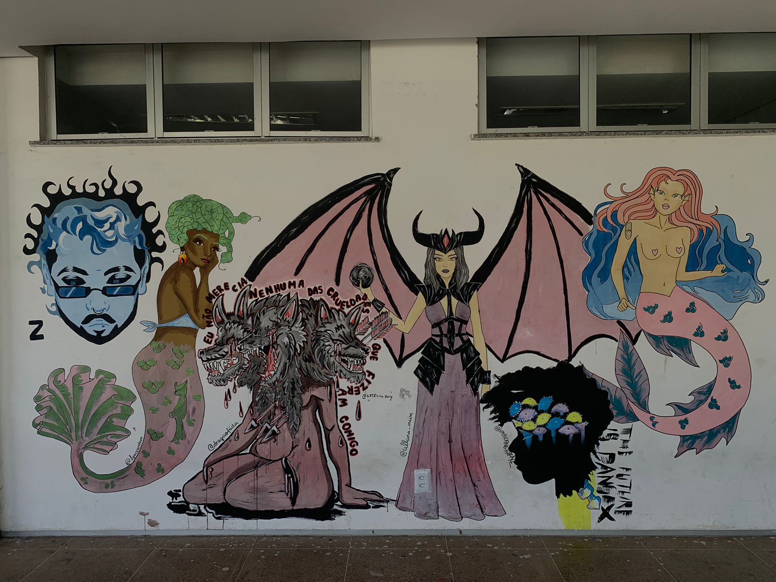Mural colorido em uma parede branca, sob duas fileiras de janelas retangulares. A arte apresenta uma série de figuras míticas e fantásticas. Da esquerda para a direita: uma figura masculina com pele azul-claro, cabelo encaracolado azul-escuro e óculos escuros, com uma cauda de sereia verde; uma sereia com cabelo verde-claro, top bege e cauda rosa-claro com escamas verdes; no centro, uma criatura humanóide com múltiplos rostos de lobo ou cão de três cabeças sobre um corpo de tom de pele rosado, sentada; ao lado, uma figura feminina com pele cinza, chifres e grandes asas rosadas, vestindo um vestido roxo e armadura preta (uma demônia ou súcubo); e, mais à direita, uma sereia de cabelo rosa, topless e cauda rosa com escamas azuis. Na extrema direita, um perfil de cabeça negra com cabelo estilizado e peixes coloridos.