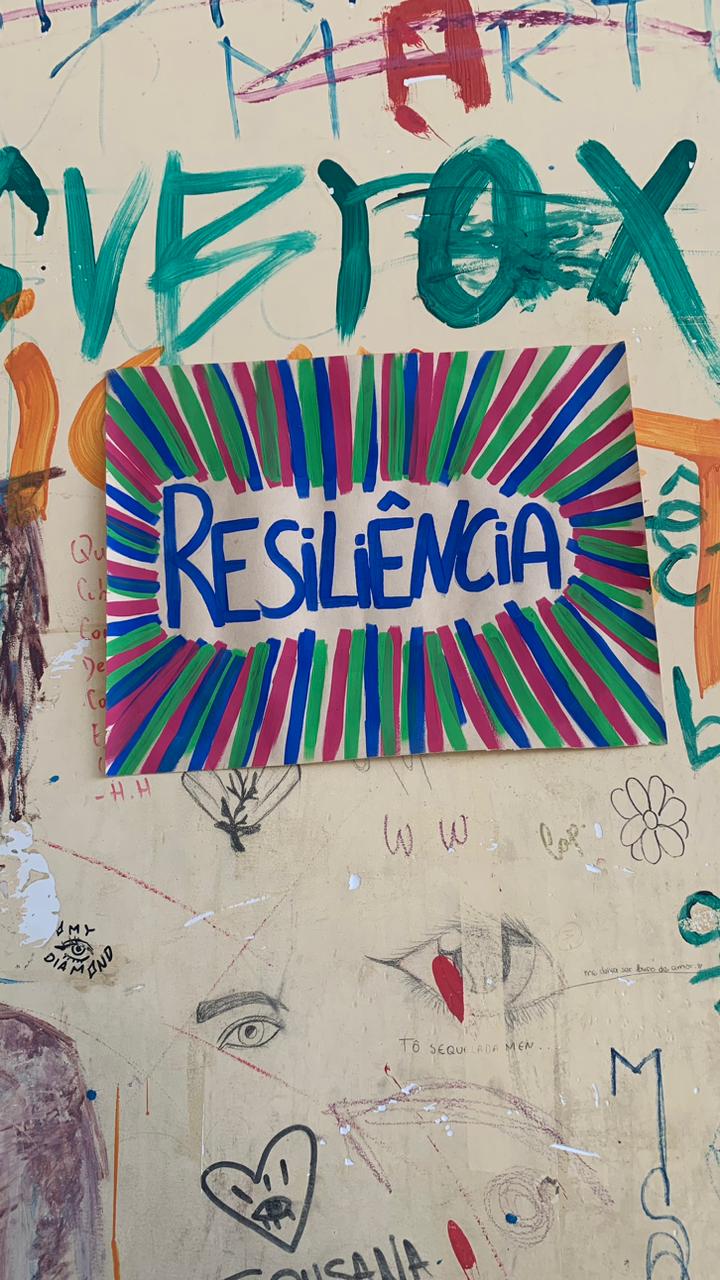 Cartaz retangular colorido afixado em uma parede amarelada e coberta de grafites. O cartaz exibe a palavra "RESILIÊNCIA" em letras maiúsculas azuis, em destaque no centro, cercada por um padrão de raios de sol em cores vibrantes, alternando entre azul, verde e magenta.
