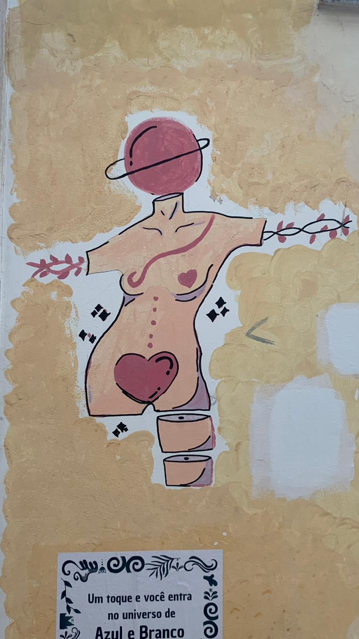 Desenho estilizado pintado em uma parede de cor amarelada, parcialmente descascada, que representa um corpo feminino. No lugar da cabeça, há um planeta com um anel (similar a Saturno), pintado em tons de rosa-avermelhado. O corpo é delineado em preto com uma cor de pele pêssego, apresentando traços de feminilidade, incluindo um coração rosa-escuro no abdômen inferior e outro menor no peito. O corpo tem galhos de folhas que se estendem dos braços e um laço rosa-avermelhado que cruza o tronco. A parte inferior do corpo é cortada e substituída por três formas cilíndricas empilhadas, possivelmente representando baldes ou tambores. A figura é ladeada por pequenos símbolos gráficos pretos.