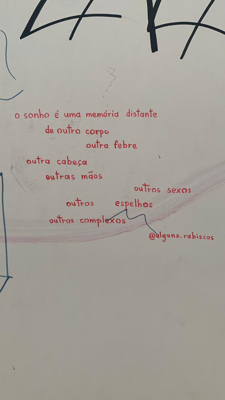 Poema escrito à mão em tinta vermelha em uma parede branca, sob um grande grafite preto e azul parcialmente visível no topo. O poema, disposto em estrofes curtas, diz: "o sonho é uma memória distante de outro corpo outra febre outra cabeça outras mãos outras outras complexos outros sexos espelhos"