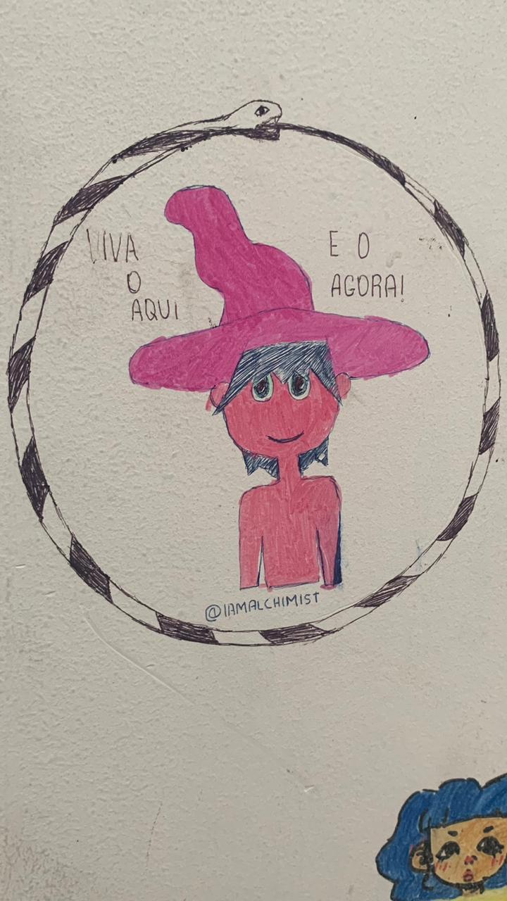 O desenho principal é um círculo que forma um Ouroboros (uma cobra mordendo a própria cauda), feito com listras pretas e brancas. Dentro do círculo, há um desenho simples de uma pessoa (ou personagem) com o rosto e corpo em tom de rosa-avermelhado, cabelo azul-escuro e um grande chapéu pontudo na cor rosa-choque, com um sorriso no rosto. Escrito em letras finas dentro do círculo, nas laterais do personagem, estão as frases "VIVA O AQUI" e "E O AGORA!".