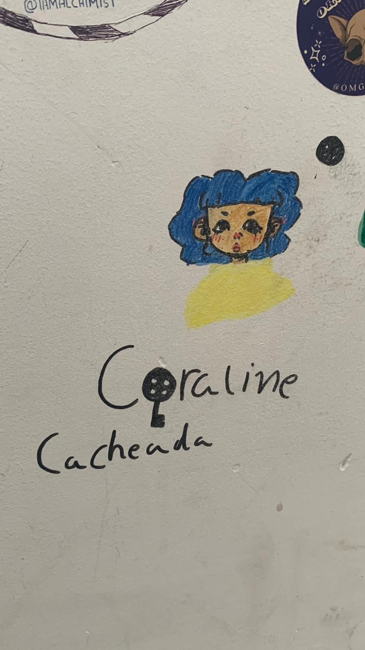 Mural em parede branca com um desenho da personagem Coraline. O desenho, no centro superior, apresenta a personagem com cabelo azul encaracolado e uma blusa amarela, com um estilo de olhos expressivo. Abaixo, a frase "Coraline Cacheada" está escrita em caneta preta; a letra 'O' na palavra "Coraline" é substituída por um desenho da chave com cabeça de botão, elemento característico da personagem.