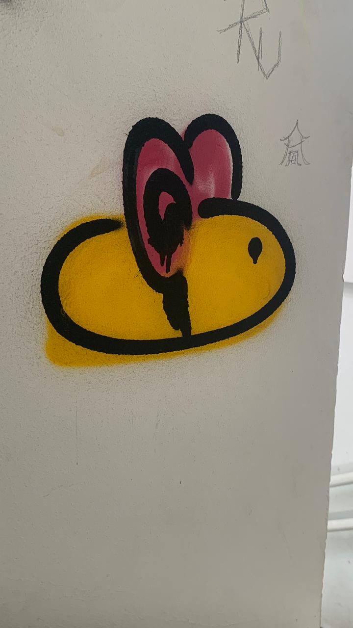 Desenho estilizado de uma abelha grafitado em uma parede branca. O corpo oval é preenchido com amarelo vibrante e contornado com spray preto grosso. Uma das asas, localizada na parte superior, é em formato de coração e é preenchida com rosa-choque, com um espiral preto no centro. O desenho tem um pequeno olho preto na frente.